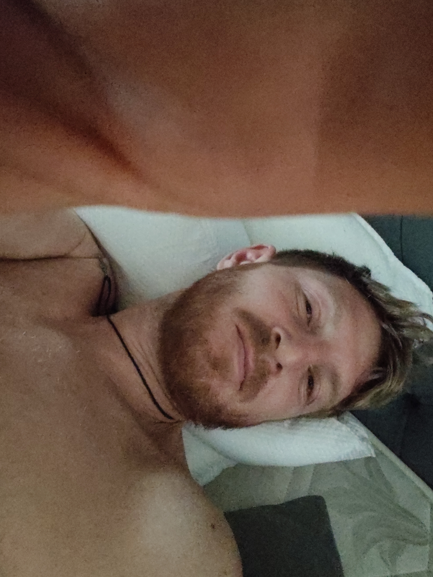 The image is a selfie taken by a man with light skin, reddish-brown hair, and a beard. He is lying down on a pillow, with his head tilted to the side. His eyes are open, and he appears to be looking at the camera. The background is blurry, but it seems to be an indoor setting. The man is wearing a black necklace. The lighting in the image is dim, casting shadows on his face.