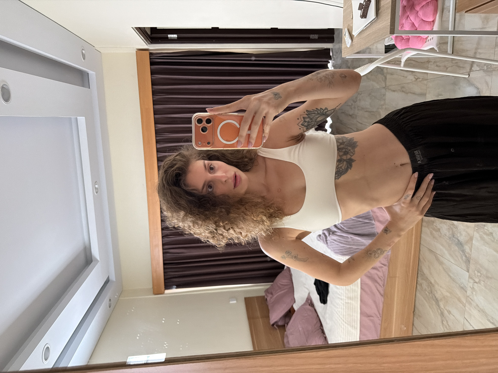 A mirror selfie captures a young woman in an indoor setting, likely a bedroom or studio apartment. The image is oriented vertically, showing the woman's reflection from approximately the waist up. She has curly brown hair and a neutral expression, looking directly into the camera. She wears a white sports bra or crop top and black loose-fitting bottoms. Tattoos are visible on both of her arms, including a butterfly design on her right hand, a floral design on her left arm, and another large floral/mandala-like tattoo on her left ribcage. Her fingernails are painted a light pink. She holds a smartphone with an orange case featuring a prominent white circular Magsafe ring.
The background shows a room with light-colored tiled flooring and a bed with purple and white bedding and a wooden headboard. A window with dark brown blinds or curtains is visible behind the woman. To her right, a small light-colored table holds various items, including a pink fluffy object, a hair clip, and what appears to be a remote control. Above, a modern white ceiling light fixture is partially visible in the mirror's reflection. Natural light illuminates the scene, suggesting it is daytime. On the waistband of her black bottoms, faint white text reads "AJUM" (or "AYUM") with "CIRCULAR" above it, likely a brand name. The overall impression is casual and personal, typical of a selfie taken at home.