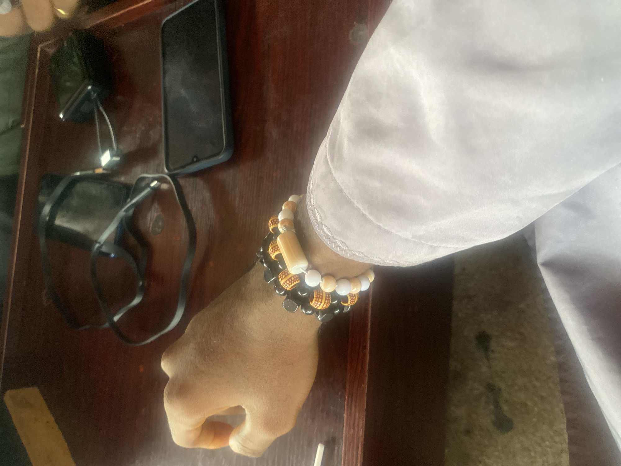 The scene depicts a first-person view of a dark wooden table in Howayi, Nigeria. A person's right forearm, with medium-brown skin, rests on the left side of the frame. Three multi-strand beaded bracelets adorn the wrist: one predominantly black, one featuring black beads interspersed with patterned orange/yellow barrel beads and a central cream rectangular bead, and one composed of cream and light orange/beige round beads. The individual wears a light gray or off-white long-sleeved garment, with a visible stitched cuff at the lower right. On the table, a black smartphone in a dark case lies in the mid-right, alongside a black portable charger connected via a light-colored USB-C cable to an obscured device. Several tangled black cables are visible in the upper-mid section. A light brown wooden object is present in the upper left background, and a small, light-colored object is partially visible at the bottom left edge. The setting is indoors.