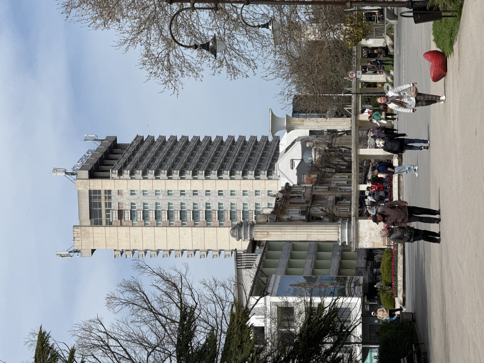 The image captures a street scene in Varna, Bulgaria. A tall, Soviet-era hotel building dominates the background. A public pedestrian area is bustling with people of varying ages and attires. Some are walking, others standing, and a few are seated on benches. Along the walkway, there are decorative elements like streetlights and sculptures, including a large red heart. Trees are visible, some bare-branched and some evergreen. The sky is partly cloudy.