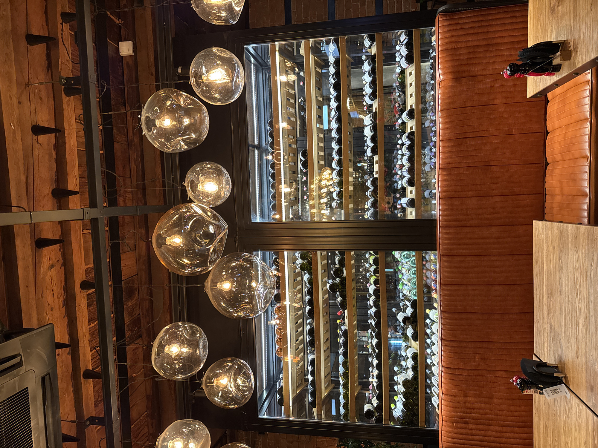 The image shows the interior of a restaurant or bar, featuring a large wine rack filled with numerous wine bottles. Adjacent to the wine rack is a booth with orange, horizontally-stitched upholstery. On the table next to the booth, there's a black wallet or small bag. Above, a complex lighting fixture consisting of multiple glass orbs hangs, casting a warm glow. The ceiling and walls in the background suggest a rustic interior design with exposed brick or wood. The location is Unknown, Unknown.