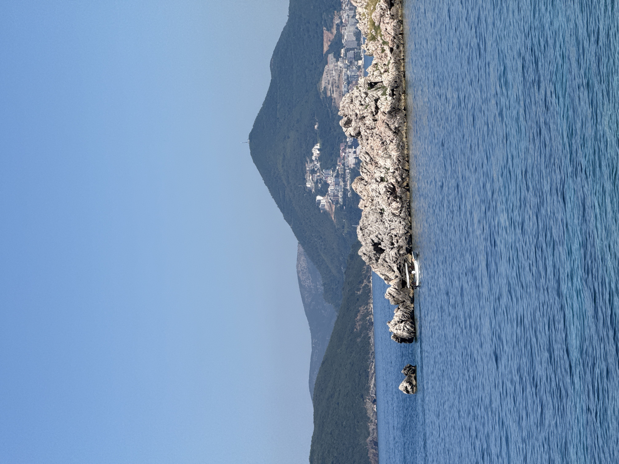 A scenic view of the coastline near Sveti Stefan, Montenegro, unfolds under a clear blue sky. The foreground is dominated by the vast expanse of the Adriatic Sea, its surface rippling gently. To the right, a cluster of rocky outcrops juts out of the water, providing a rugged contrast to the calm sea. A small white boat with a canopy is moored near these rocks, with a person visible onboard. In the mid-ground, rolling green hills rise from the water's edge, their slopes covered in dense vegetation. Further in the background, a larger mountain looms, its peak adorned with a communication tower. White buildings are scattered across the mountainside, suggesting a settlement or resort. The overall scene captures the natural beauty and coastal development of the area.