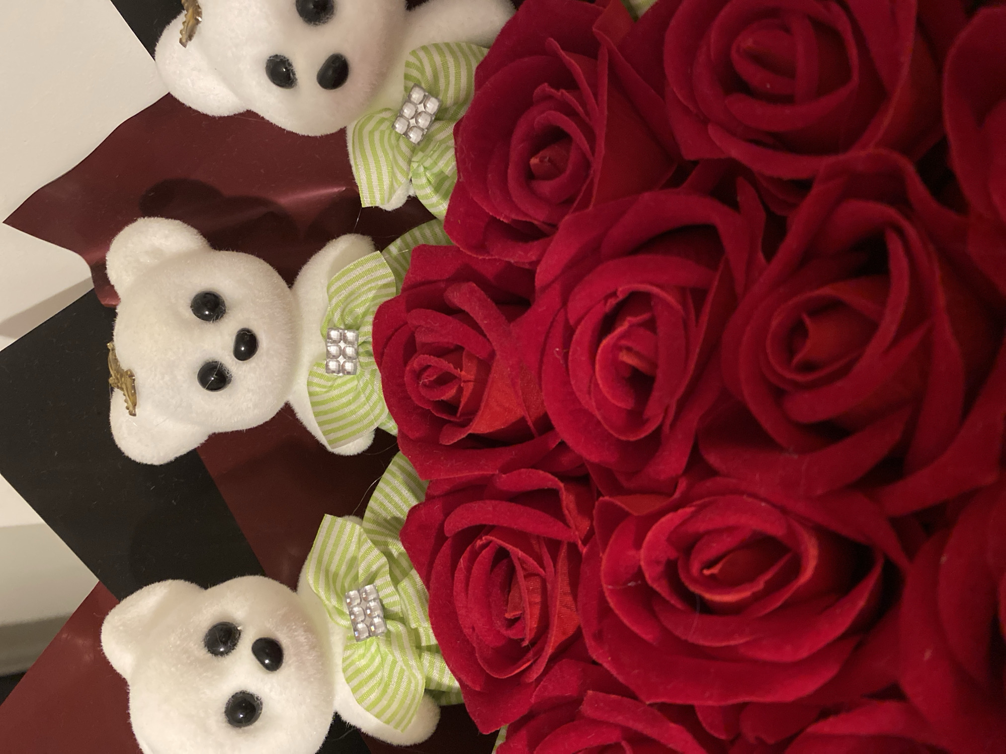 A close-up, overhead shot displays a bouquet. The arrangement features multiple red roses clustered together, taking up the majority of the right side of the frame. To the left, three small, white teddy bears are positioned in a row, each adorned with a green and white striped bow featuring a jeweled accent. The background is a striped, maroon-and-white surface. The environment cannot be determined from the image. There is no evidence to determine the location is in the city of Alanya, Turkey.