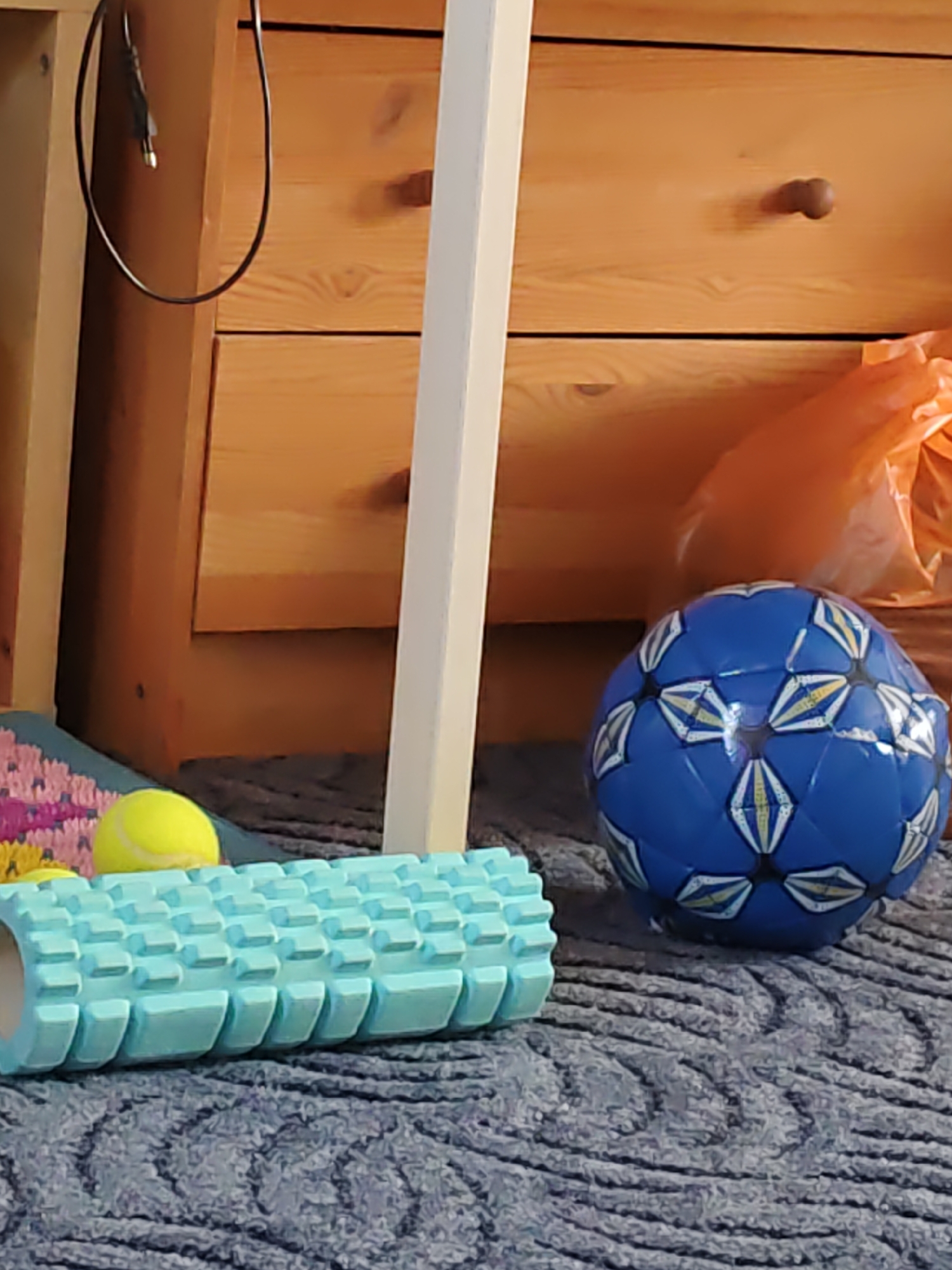 The image depicts an indoor scene, likely a bedroom or living area, with objects associated with physical activity and storage. In the foreground, a light blue foam roller lies horizontally on a dark grey carpet with a subtle textured pattern. To the right of the foam roller, a blue soccer ball with white and yellow hexagonal patterns is visible. Behind the soccer ball, a crumpled orange plastic bag is partially visible. In the background, a wooden dresser with two drawers and a single round knob is present. A black cable with a metal connector is draped over the side of the dresser. A small yellow tennis ball is situated to the left of the foam roller, resting on a multicolored woven mat. The overall lighting suggests an indoor setting, possibly during the day with ambient light. The location is identified as Rostov-on-Don, Russia. There are no people or animals visible in the image.