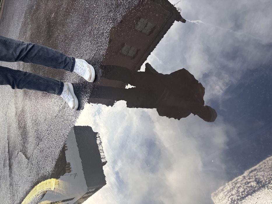This image depicts an urban street scene in the city of London, United Kingdom, captured from an overhead perspective. The lower body of an individual is prominently featured, wearing dark blue jeans and white sneakers with light beige trim. The person stands on dark, wet asphalt pavement. A bright yellow painted line is visible on the asphalt to the immediate left of the individual's left foot, partially submerged by a large, reflective puddle. The puddle occupies the lower half of the frame and reflects elements of the surrounding environment. Reflected in the puddle are: the dark, silhouetted upper body and head of the standing individual; a dark, flat-roofed building to the left of the individual's reflection; a reddish-brown brick building with multiple rectangular windows to the right of the individual's reflection; and a partly cloudy sky displaying white clouds against a pale blue backdrop. The wet ground conditions indicate recent precipitation.