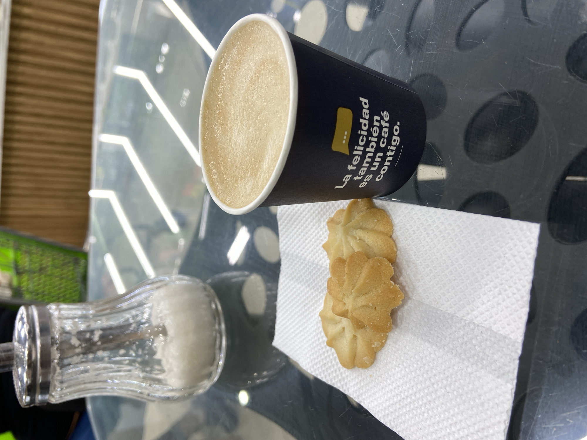 A close-up, eye-level shot shows a cafe table setting in Juan Griego, Venezuela. In the foreground, two star-shaped cookies rest on a white paper napkin. To the right, a black paper cup filled with a frothy, light brown beverage, presumably coffee, is positioned. The cup has white text in Spanish: "La felicidad también es un café contigo." Behind the cookies and coffee cup, a clear glass sugar dispenser with a silver lid is visible, containing white granulated sugar. The table surface is black and reflective, showing faint circular patterns and light streaks from overhead lighting. In the blurred background, a green wire basket and a wooden partition are discernible, suggesting an indoor or covered outdoor seating area. The overall ambiance is that of a relaxed cafe experience.