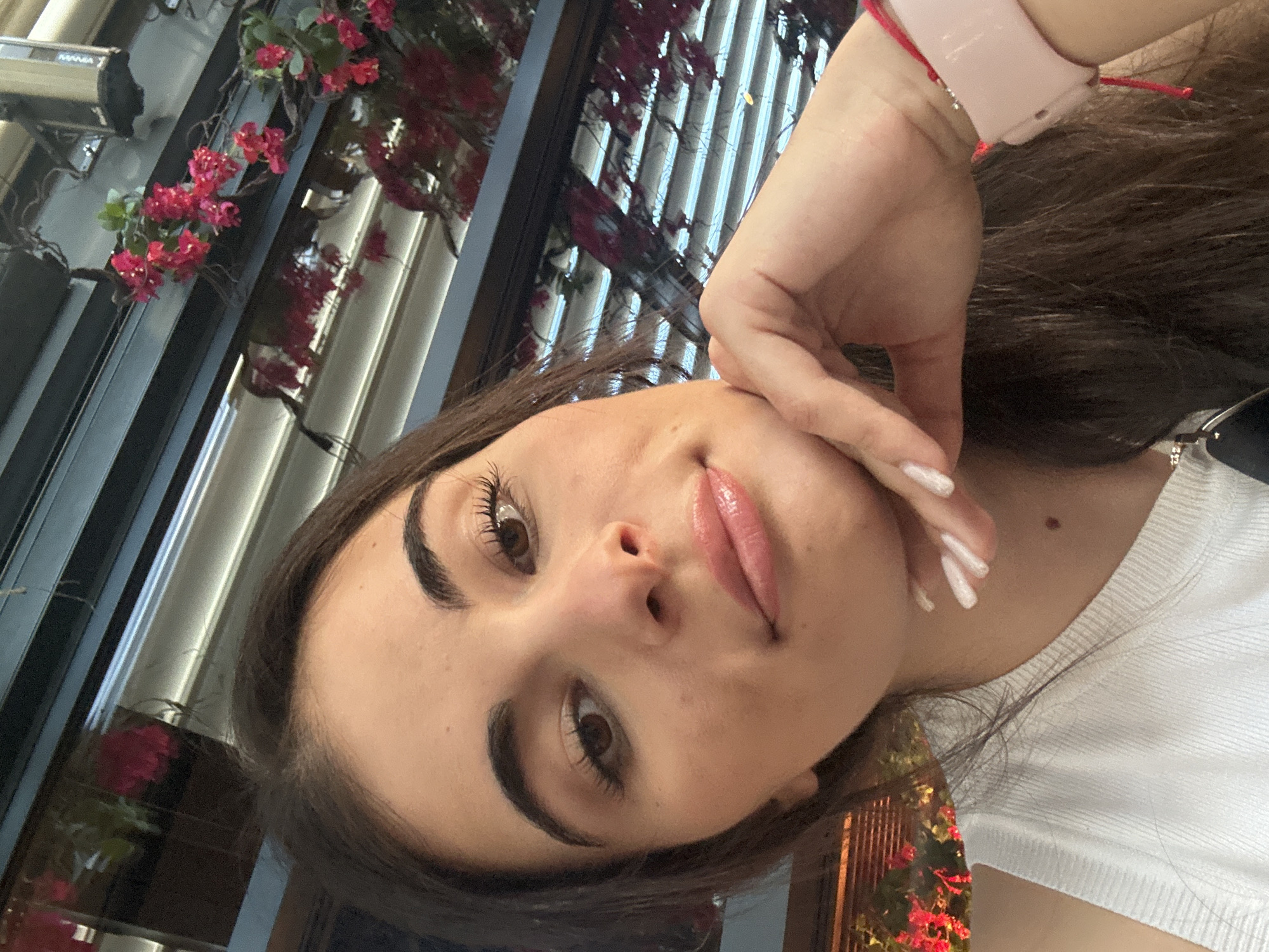 A young woman with long, dark brown hair is captured in a close-up selfie. She is resting her chin on her hand, her fingers adorned with white nail polish. She is wearing a white tank top, and a pink smartwatch with a red string is visible on her wrist. Behind her, there is a dark horizontal structure with blinds, and further back, vibrant pink flowers are blooming, suggesting an outdoor or semi-outdoor setting. The lighting suggests it may be daytime.