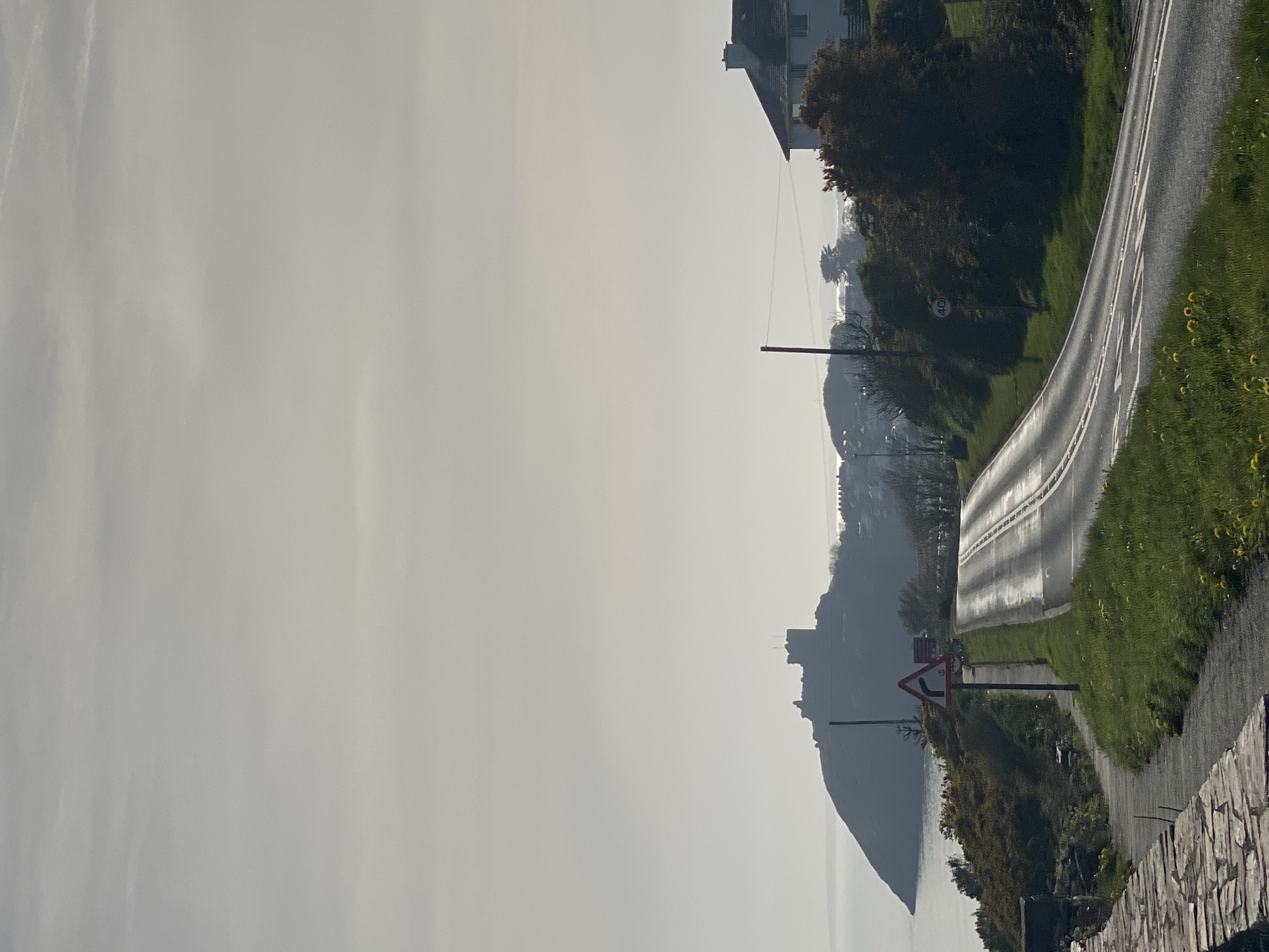 The image depicts a coastal scene in Criccieth, United Kingdom, characterized by hazy conditions obscuring the distant features. A wet, winding road dominates the foreground, flanked by green grass and sporadically blooming yellow flowers. To the right, a stone wall is visible along the roadside. A triangular warning sign is present near the road. On the left, in the distance, stands Criccieth Castle, its details softened by the haze. A small section of a building is visible at the upper right corner.