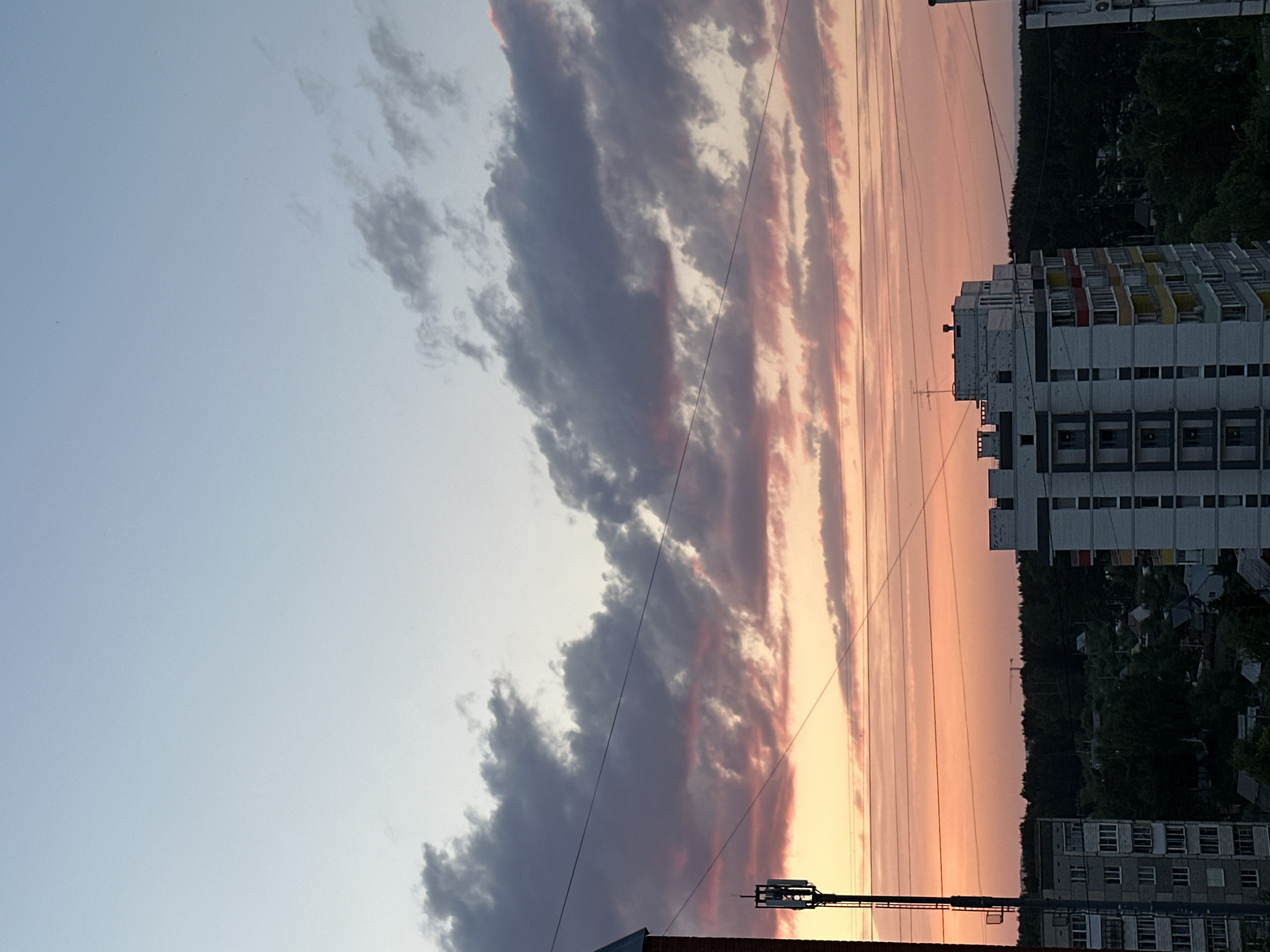 The image depicts a sky at sunset in what is likely the city of Chaikovsky, Russia. The sky shows a gradient from a light blue at the top left to a pinkish-orange on the right, with long bands of grey and pink clouds aligned vertically in the center of the image. There are power lines visible in the foreground, running vertically across the clouds. On the right side of the frame, there are multiple apartment buildings, some taller than others, suggesting an urban environment. Behind the buildings are trees. In the lower portion of the image, there are antennae and a rooftop. The overall scene suggests a residential area with urban and natural elements.