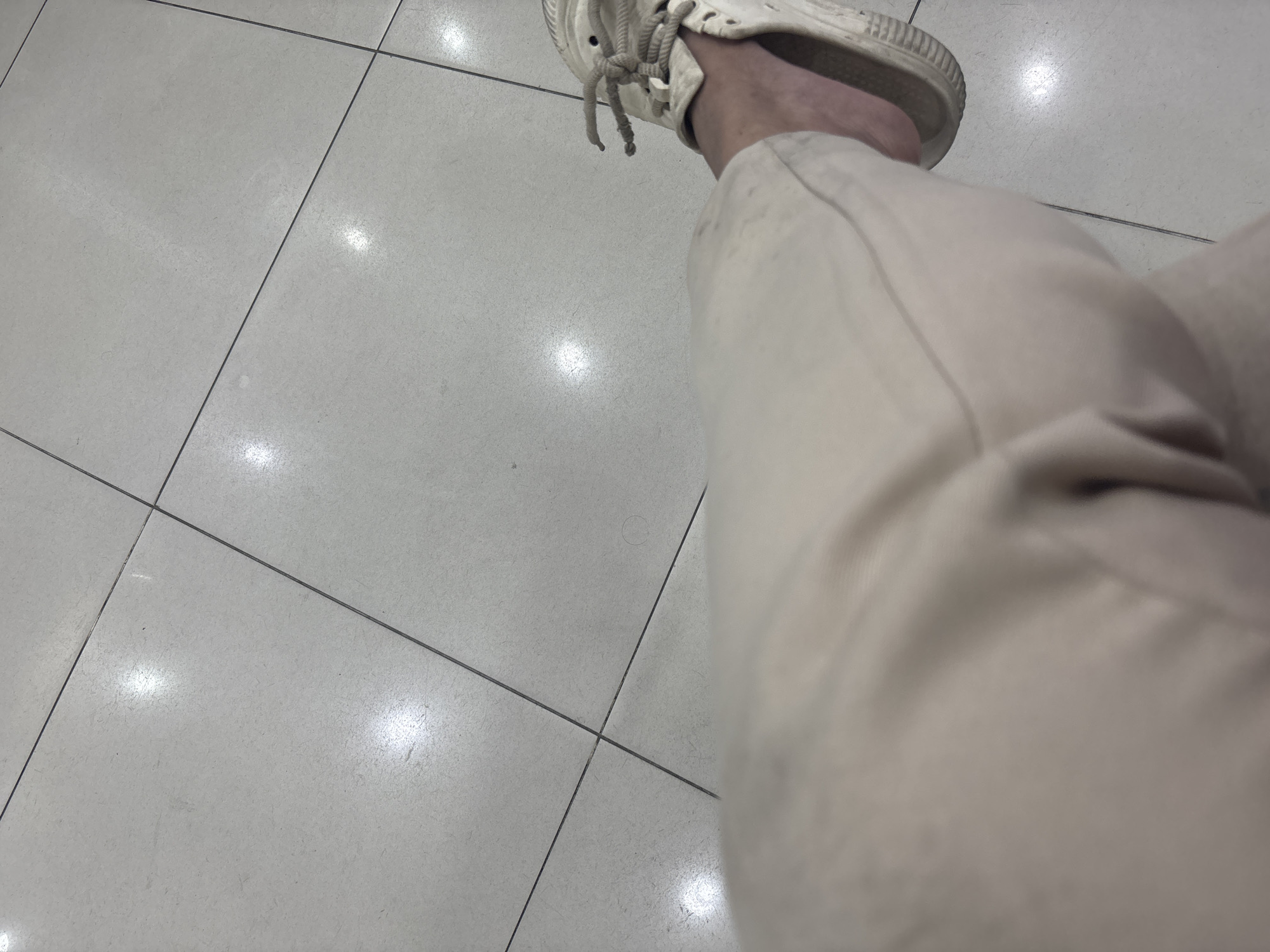 The image shows a person's legs and feet standing on a tiled floor. The floor is made of large, light-colored square tiles with dark grout lines. The lighting on the floor suggests it is indoors, possibly a commercial space or a public area. The person is wearing light-colored pants and a white sneaker. The sneaker is a low-top style with laces. The lower part of the pants and the foot are in focus, while the upper part of the pants is blurred. There are reflections of light on the tiled surface. No specific landmarks or identifying features of North Jakarta, Indonesia are visible in the image.