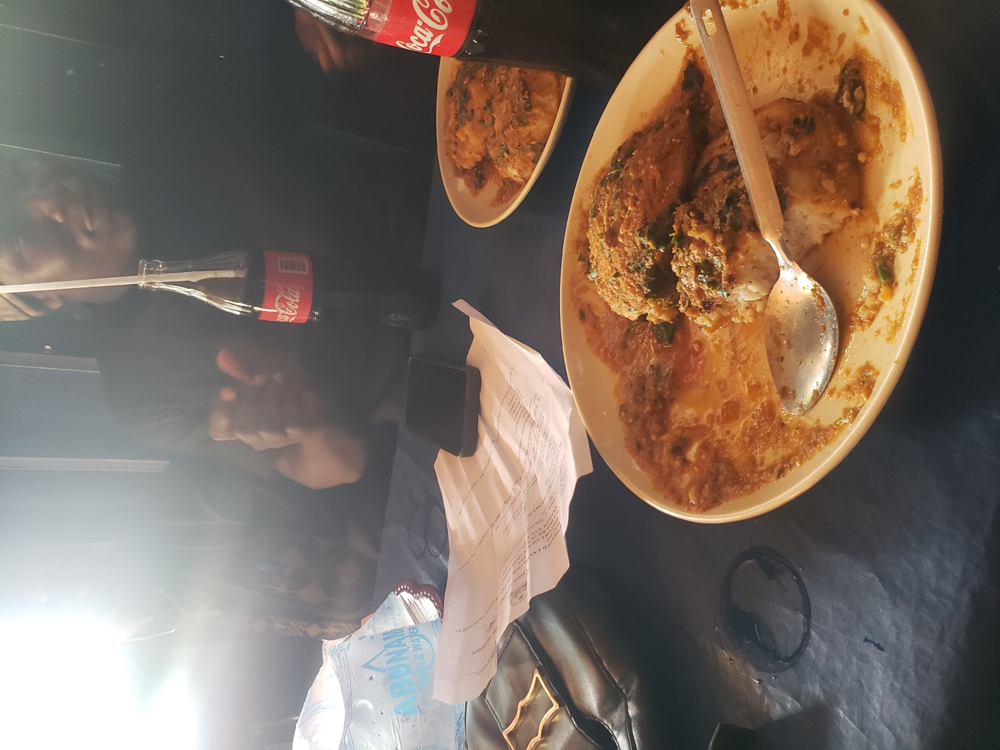 The image depicts a dimly lit dining setting, likely a restaurant or informal eatery in Kaduna, Nigeria. In the foreground, a partially eaten meal of rice topped with a savory stew or sauce is presented on a pale yellow plate. A spoon rests on the plate, having been used to consume the food. To the right, a Coca-Cola bottle stands prominently, its red label clear. Another bottle of Coca-Cola is partially visible behind it. A crumpled paper, possibly a menu or receipt, lies between the food and a dark object, which appears to be a mobile phone. To the left, a plastic bag of water, labeled "ABUNAM TABLE WATER," is visible. A black leather bag with a distinctive gold buckle is also present on the table. The overall impression is of a casual meal, possibly shared, given the presence of two plates of food visible in the frame. The lighting suggests an indoor environment, with a bright light source creating a lens flare effect in the upper left quadrant of the image.