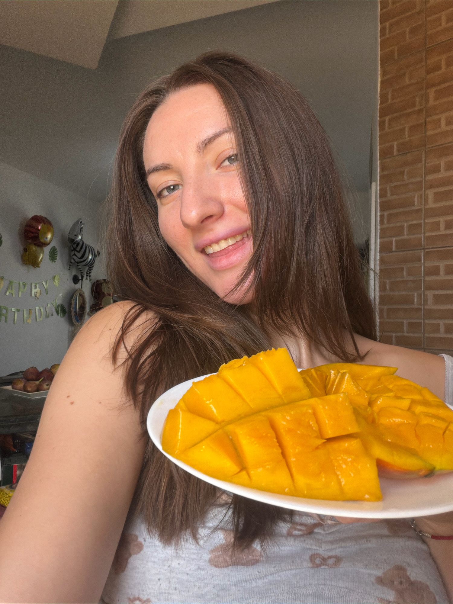 A young woman with long, brown hair is taking a selfie, holding a white plate with sliced mango. She is smiling and sticking her tongue out slightly. She is wearing a light grey t-shirt with a floral pattern. In the background, there is a wall with exposed brick, a window, and some decorations that appear to be for a birthday party, including balloons shaped like a lion and a zebra, and a banner that reads "Happy Birthday". There is also a plate of red fruits visible on a shelf. The scene appears to be indoors, possibly in a home or hotel room in Sharm El Sheikh, Egypt.