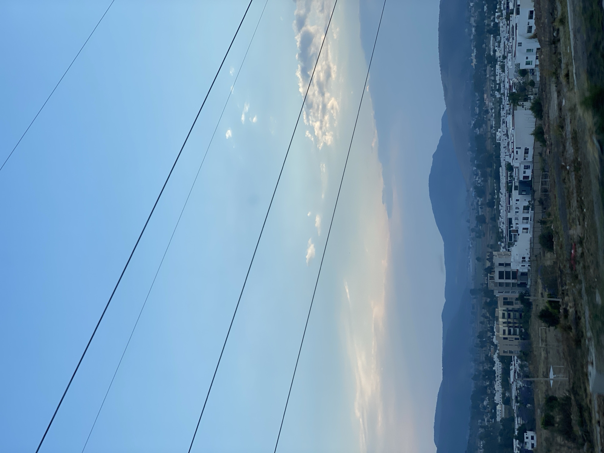 The scene shows a residential area in La Cantera, Mexico, nestled at the base of mountains. The sky is pale blue with some wispy clouds. High-tension power lines diagonally cut across the view from the left side. The mountains are silhouetted against the sky, and the residential area consists mainly of white buildings. The foreground, near the buildings, features some brown, arid vegetation.