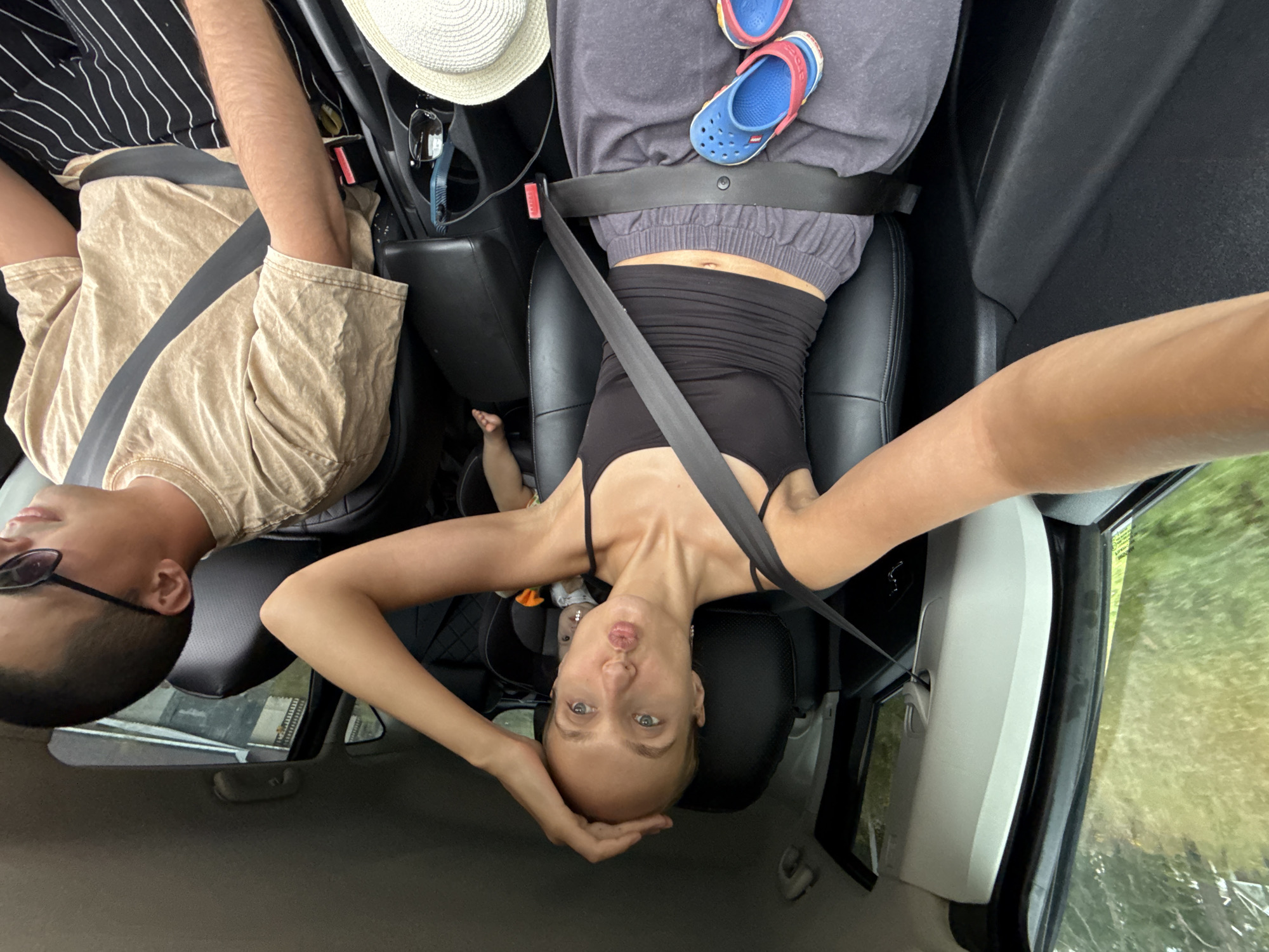 The image captures a selfie taken from inside a car in Podshivalovo, Russia. A woman in a black tank top and grey sweatpants is in the driver's seat, with a seatbelt fastened across her. She is holding her hand to her forehead and puckering her lips. In the background, a baby is visible in a car seat. To the right, a man in a tan t-shirt and striped pants is in the passenger seat, wearing sunglasses and a seatbelt. A straw hat and sunglasses are on the center console. The exterior of the car shows a blurry green, grassy landscape, suggesting the vehicle is in motion or parked near a rural area.