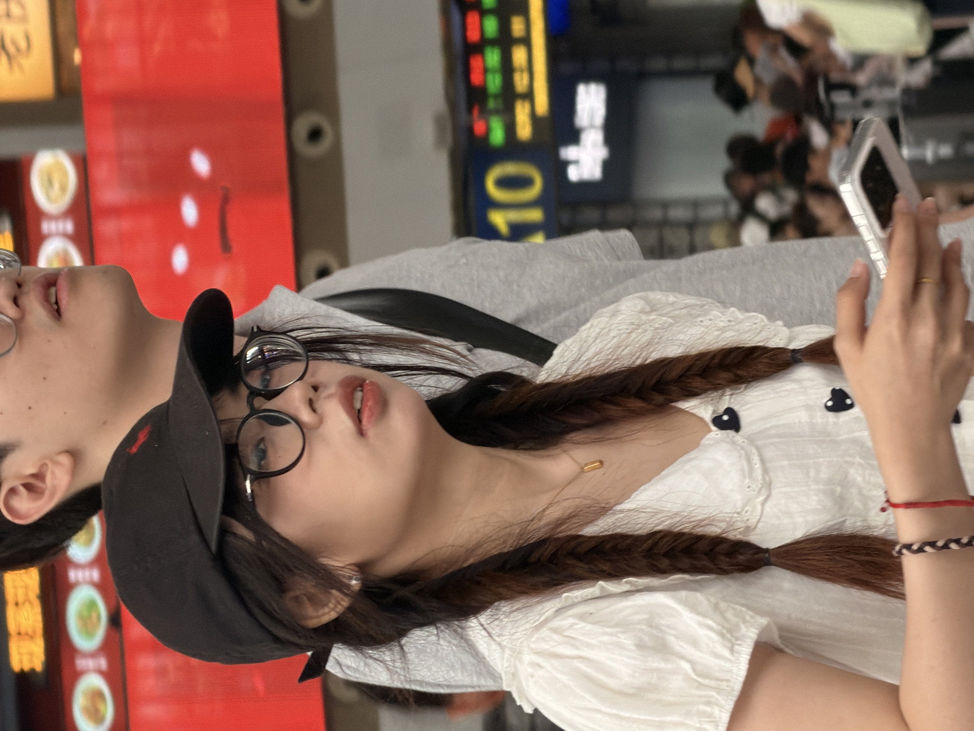 In Panyu District, China, a young woman wearing a black baseball cap and round glasses is captured in a medium shot, holding a white smartphone. She has dark hair styled in two braids and is dressed in a white ruffled top with black heart-shaped buttons. Beside her, partially visible, is another person wearing a grey hooded sweatshirt. The background suggests a public space, possibly a train station or a shopping area, with blurred figures and illuminated signs displaying Chinese characters and numbers. The lighting is ambient, casting soft shadows.