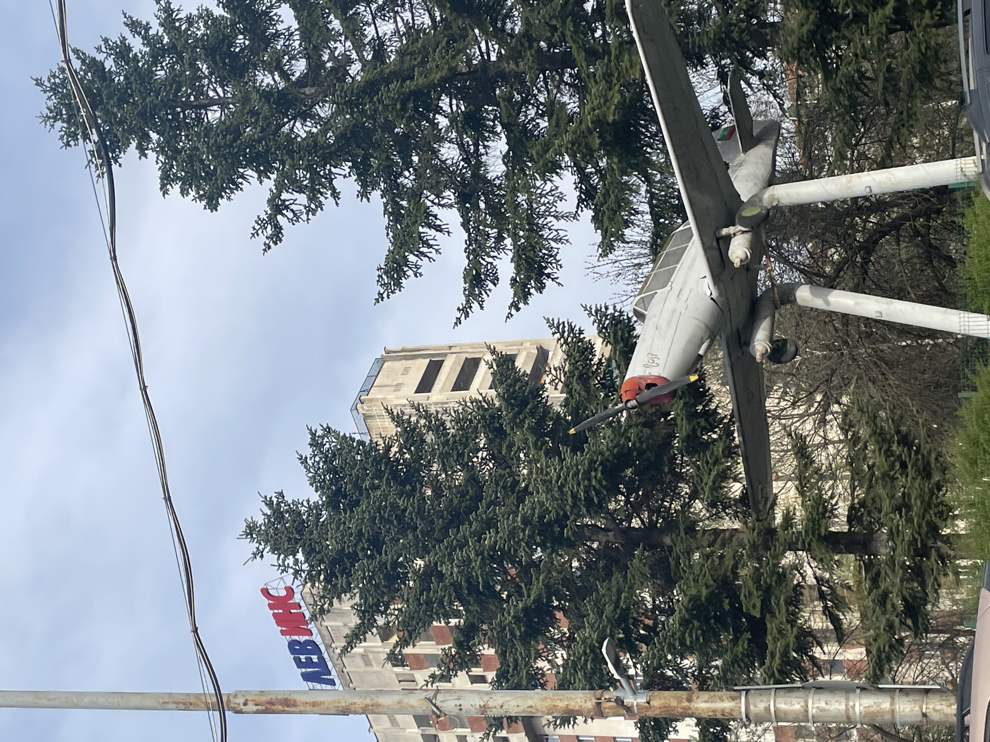 The image captures an outdoor scene in Sofia, Bulgaria, on an overcast day, featuring a static display of an old propeller airplane. The airplane, painted predominantly in light gray, has a distinct red nose cone and a single propeller. Its tail fin displays the colors of the Bulgarian flag: white, green, and red stripes. The aircraft is mounted on two prominent white, cylindrical supports, elevating it above the ground.
Dense evergreen trees, likely fir or spruce, are scattered throughout the mid-ground, their dark green foliage partially obscuring the background. Through the trees, several buildings are visible. On the left side of the frame, a building features a large sign with the Cyrillic text "ЛЕВИНС" displayed in red and blue letters, translating to "LEVINS". Further back and to the right, a tall, rectangular building with multiple dark window openings stands prominently against the cloudy sky.
In the upper left portion of the image, dark power lines or cables stretch diagonally across the light gray, overcast sky. The lower foreground shows fragments of rusty metal structures, possibly part of the airplane's support or street infrastructure. The overall scene suggests a public space, possibly a park or square, where the airplane serves as a monument or a display. No human activity or specific event is depicted; the scene is static and quiet.
