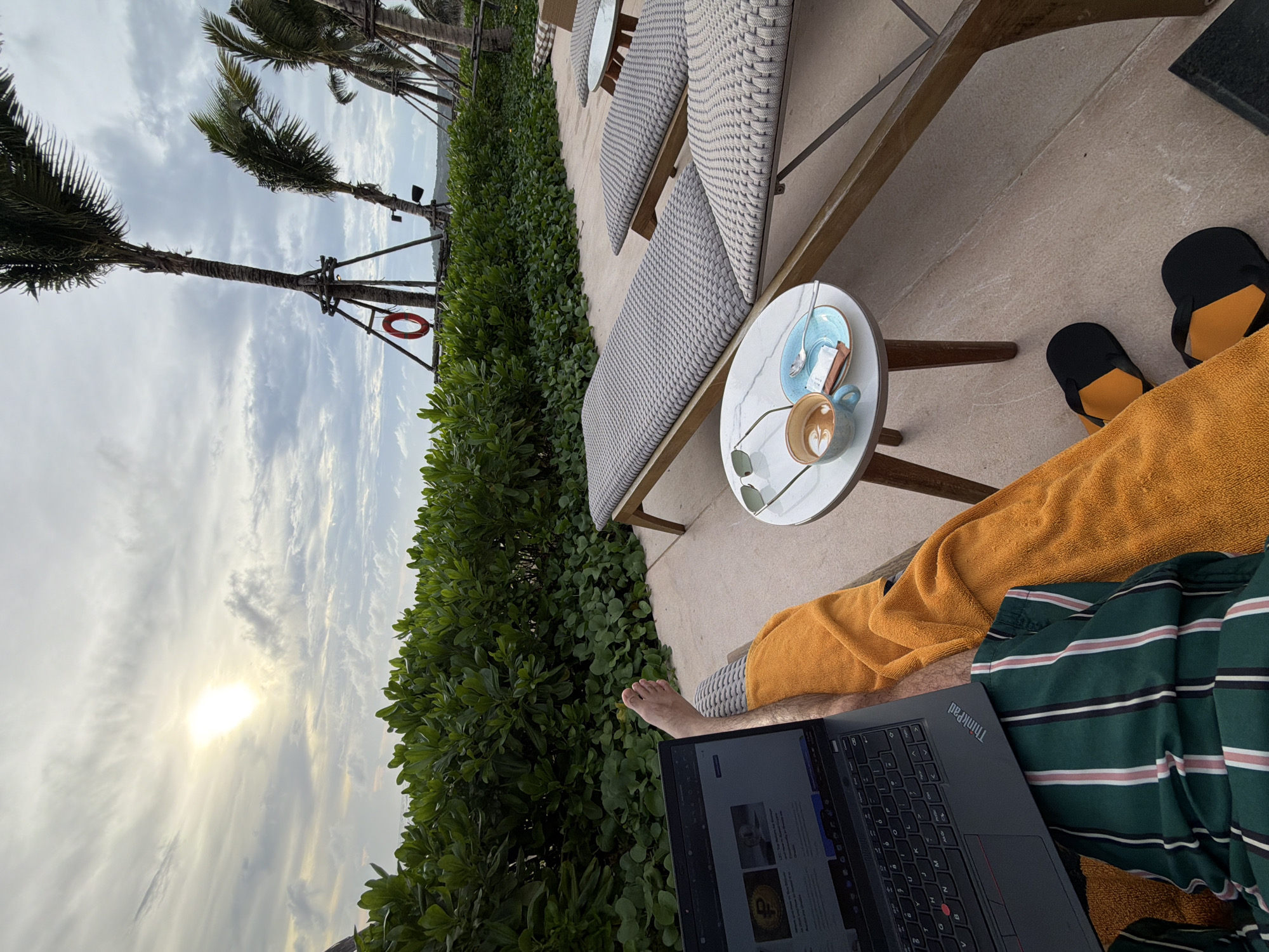 A view from a beach lounger in Choeng Thale, Thailand. A person with a laptop, wearing a green striped shirt and covered by an orange towel, is positioned in the lounger. A table next to the lounger holds a cup of coffee, sunglasses, and a dish with a spoon and sweeteners. Black and orange sandals are visible near the lounger. The lounger itself is upholstered in a grey woven pattern. Palm trees and a dense hedge line the background, with a cloudy sky and the sun visible behind the trees. A life preserver is attached to a palm tree trunk.