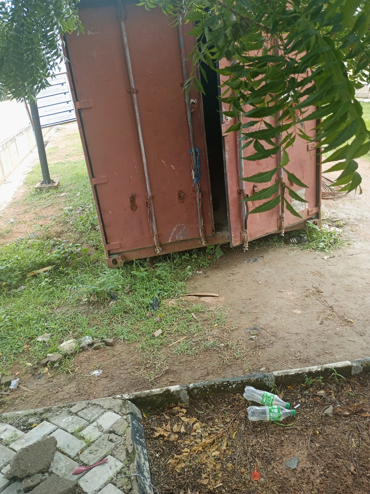 The image depicts an outdoor scene in Jalingo, Nigeria, featuring a large, rusted red shipping container placed on uneven ground. The container is open, revealing dark interior space. Its doors are secured by multiple vertical metal bars and rusty latches. A blue rope is visible hanging from one of the doors. Lush green foliage from a nearby tree hangs over the top of the container, with some leaves obscuring parts of its surface. The ground surrounding the container is a mix of dirt, sparse green grass, and debris, including scattered white plastic, what appears to be discarded black shoes, and some construction materials. In the foreground, to the right, two empty plastic bottles with green labels are lying on the ground near a concrete border. To the left, a section of a paved walkway made of grey interlocking bricks is visible, with broken pieces of concrete and a red plastic spoon on top. A metal pole supporting a sign is also visible in the background. The overall impression is one of an urban or semi-urban outdoor space, possibly a yard or an undeveloped area, with a sense of disuse or neglect.