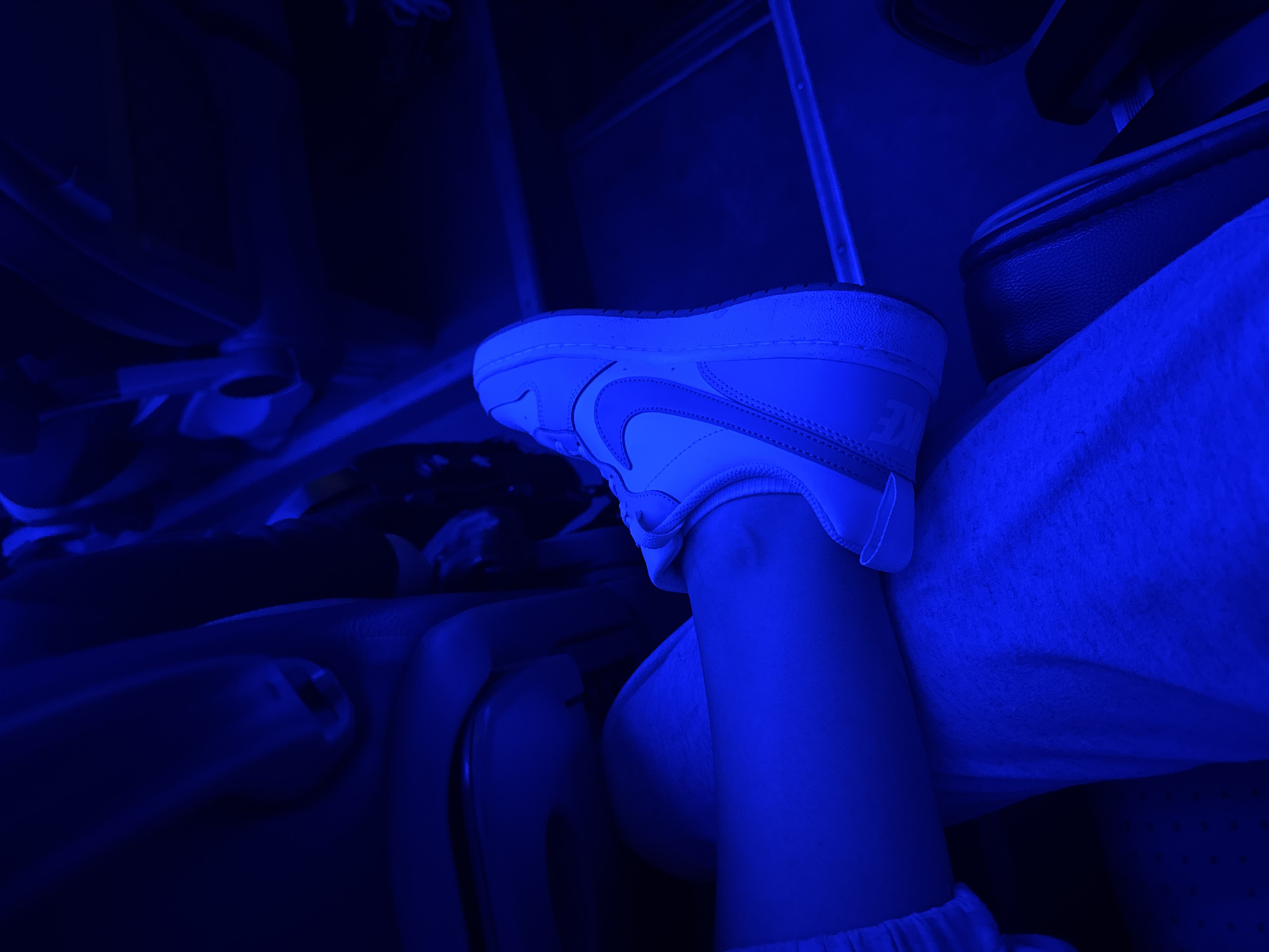 The image depicts a close-up view of a person's lower legs and feet, adorned with white Nike sneakers. The scene is bathed in a strong blue light, which casts deep shadows and highlights the textures of the clothing and footwear. The person appears to be seated, possibly on a bus or train, with the background suggesting an interior of a vehicle. The blue lighting creates a dramatic and somewhat surreal atmosphere, obscuring finer details of the surroundings. The location is specified as Podgornyi Driush, Russia.