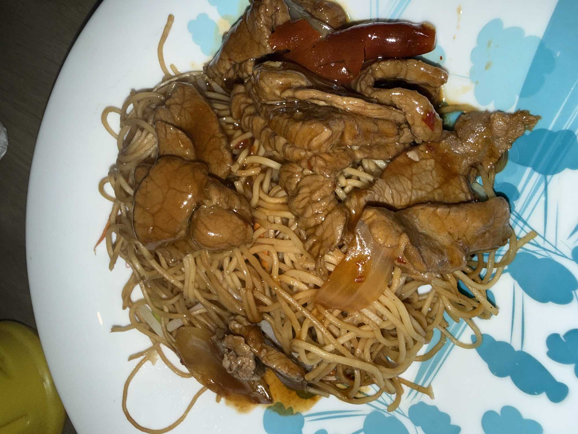 A plate of food consisting of noodles and meat. The noodles appear to be thin and yellowish-brown in color, possibly egg noodles. The meat is cut into bite-sized pieces and coated in a dark sauce. The plate is white with a blue floral pattern on one side. The lighting suggests an indoor setting. There is a glimpse of what might be a yellow object to the side of the plate. The exact location is unknown.