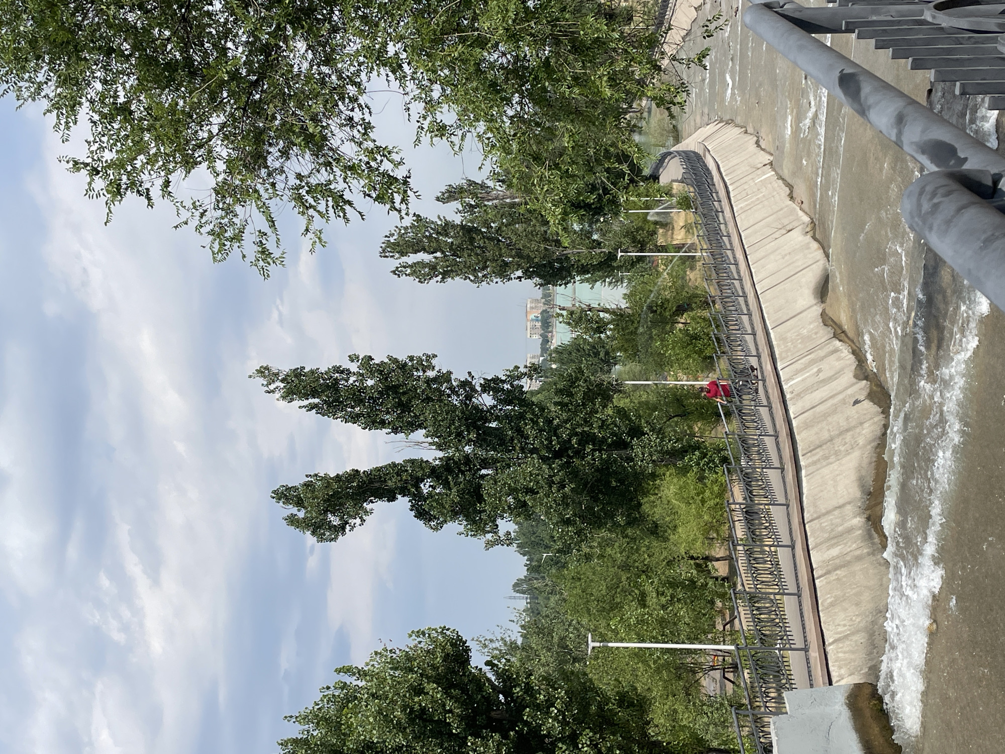 The image shows a view of the Bolshaya Almatinka River in Almaty, Kazakhstan. The river flows through a channel with concrete embankments. A walkway with a decorative metal railing runs along the embankment on the left side of the river. A person wearing a red shirt is cycling on the walkway. Trees line the walkway and the surrounding area. In the background, there are more trees and buildings, with a hazy sky overhead. The river water appears murky and is flowing rapidly with some whitewater.