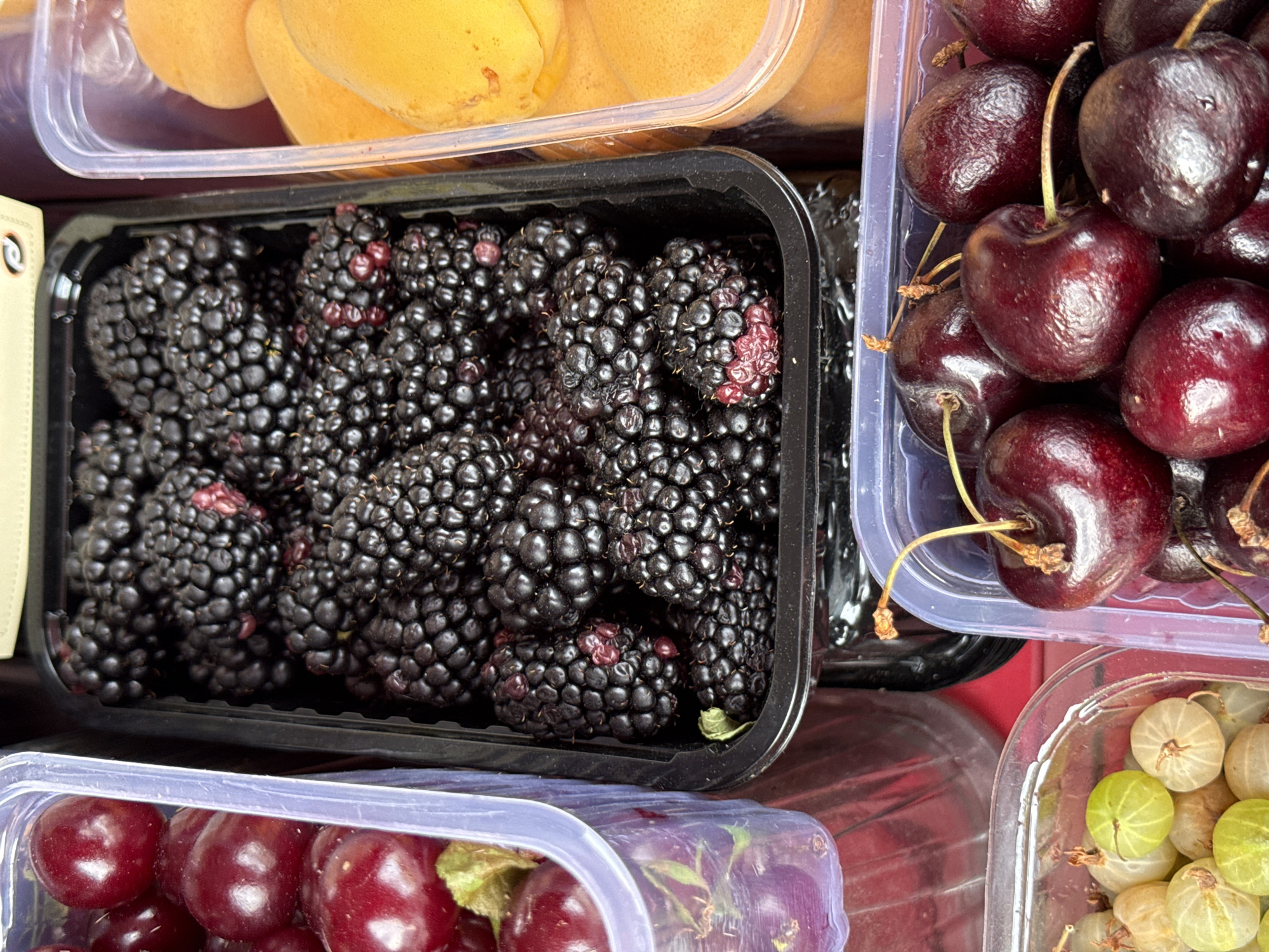 The image displays an assortment of fresh fruits in clear plastic containers. In the foreground, on the right, is a container of dark red cherries with their stems still attached. To the left of the cherries, another container holds pale green and yellow gooseberries. In the center of the image, a black plastic container is filled with ripe blackberries, some showing hints of red. To the left of the blackberries, a clear plastic container is filled with dark red cherries. On the far right, a clear plastic container holds sliced yellow apricots. The background shows indistinct shapes and colors, suggesting a market stall or grocery store setting. The lighting appears to be natural, possibly from overhead. There are no people or visible actions in the image. The context of Moscow, Russia, is not discernible from the visual elements of the photograph.