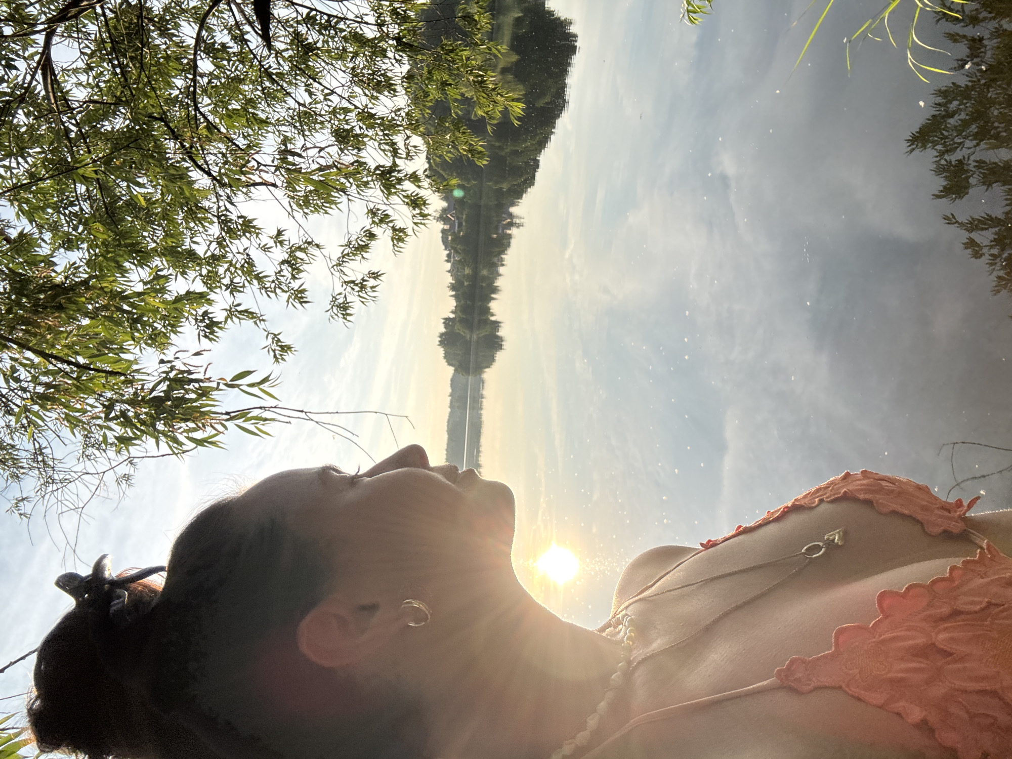 The image shows a woman in a coral-colored bikini looking out at a calm lake during sunset. The sun's glare reflects off the water, creating a shimmering effect. Trees and houses are visible on the opposite shore, and their reflections are mirrored in the lake. The woman has her hair pulled up in a bun and is wearing a necklace and hoop earrings. The scene is set against a backdrop of a clear sky with some clouds. The location is the city of Kamysh, Russia.