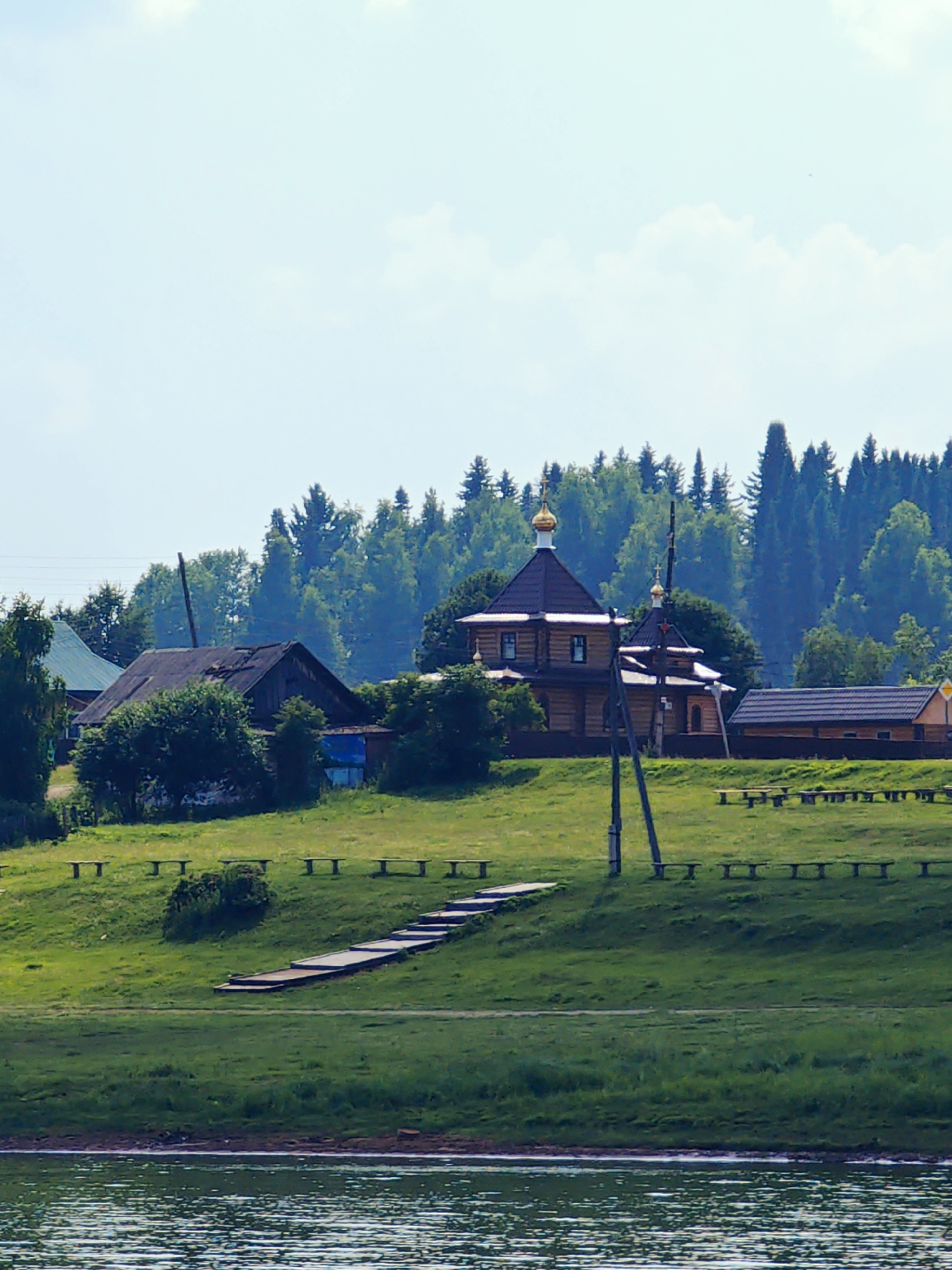 The image depicts a serene summer landscape on the outskirts of Ust-Kem, Russia, likely near a body of water, possibly the Kem River. In the foreground, the rippling surface of the water reflects the light. Beyond the water, a grassy embankment rises, dotted with numerous wooden benches arranged in rows along its crest. A wooden walkway with steps ascends the embankment, leading towards a cluster of buildings.
Dominating the mid-ground is a wooden Orthodox church with a dark brown roof and golden cupolas, its exterior constructed from light-colored logs. Adjacent to the church, to its right, is a smaller wooden structure with a dark roof, possibly a chapel or auxiliary building. Further to the left, an older, dilapidated wooden house with a dark, weathered roof stands partially obscured by lush green foliage. A tall wooden utility pole stands prominently in front of the church.
The background is characterized by a dense forest of coniferous trees, their dark green canopies creating a stark contrast against the pale blue sky, which is sparsely populated with white clouds. The overall impression is one of a peaceful rural setting, bathed in the soft light of a summer day. There are no people visible in the image.