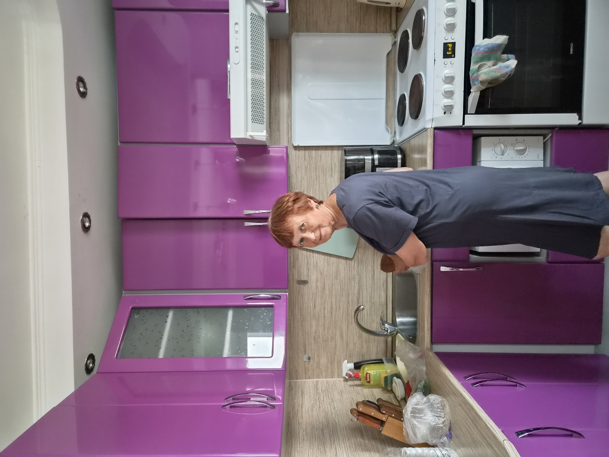 A woman with short red hair and wearing a dark grey dress stands in a kitchen. She is facing the camera and looking slightly to her left. Her right hand is holding a brown object near her chest. The kitchen has purple cabinets above and below the counter. A sink with a faucet is visible to the woman's left. To the right of the sink are cleaning supplies, including a bottle of Sanita dish soap and a spray bottle. A wooden knife block with knives is also on the counter. A coffee maker is visible behind the woman. A white stove with four burners is to the right of the woman, and an oven door with a striped dishtowel hanging from it is below the burners. A microwave is located under the counter to the left of the stove. The walls are tiled with a light beige textured pattern. The ceiling has recessed lighting fixtures. The overall environment suggests a domestic kitchen setting, likely a residential apartment. The image does not provide specific information about the city of Vladivostok, Russia.
