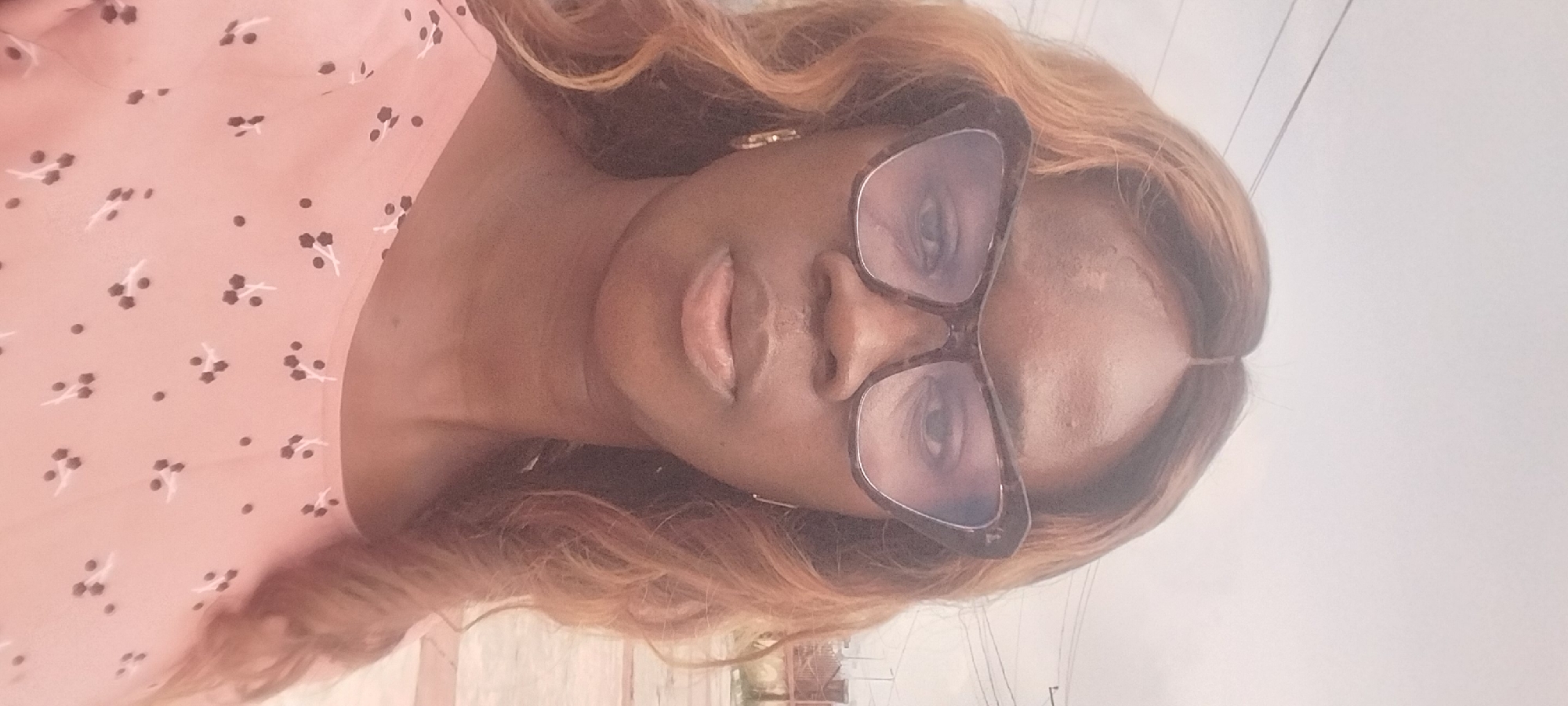 A medium shot, selfie-style photograph shows a Black woman in her late teens or early twenties looking directly at the camera. She has light brown, wavy hair that is parted in the middle and falls past her shoulders. She is wearing a pair of large, cat-eye glasses with dark brown tortoiseshell frames. Her skin is a medium brown. She is wearing a light pink shirt with a pattern of small dark floral shapes and dots. She is wearing gold hoop earrings. The background is out of focus, but appears to be outdoors in a city. There are utility wires visible in the upper portion of the background, and a fence or wall with some vegetation can be seen in the upper right. This photograph was taken in Onitsha, Nigeria.
