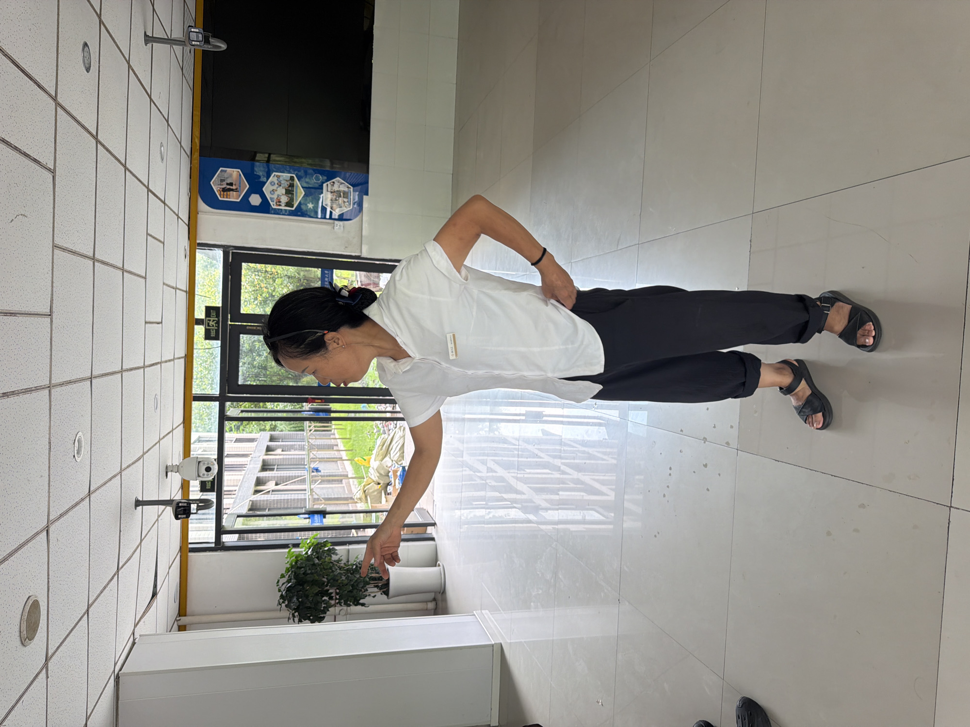 The image shows a woman standing in what appears to be an indoor hallway or entrance of a building. She is wearing a white short-sleeved collared shirt and black pants. Her left hand is pointing towards the left side of the frame, while her right hand rests on her hip. She is wearing black sandals. The environment features a tiled floor, a white wall on the left, and large glass doors and windows in the background that look out onto an outdoor area with greenery and a building under construction or renovation. There are security cameras mounted on the ceiling. To the right of the woman, there is a large dark screen or wall. The overall impression is of a functional, potentially public or commercial space. The image does not contain enough specific visual cues to determine its precise location within Jiangjin, China.
