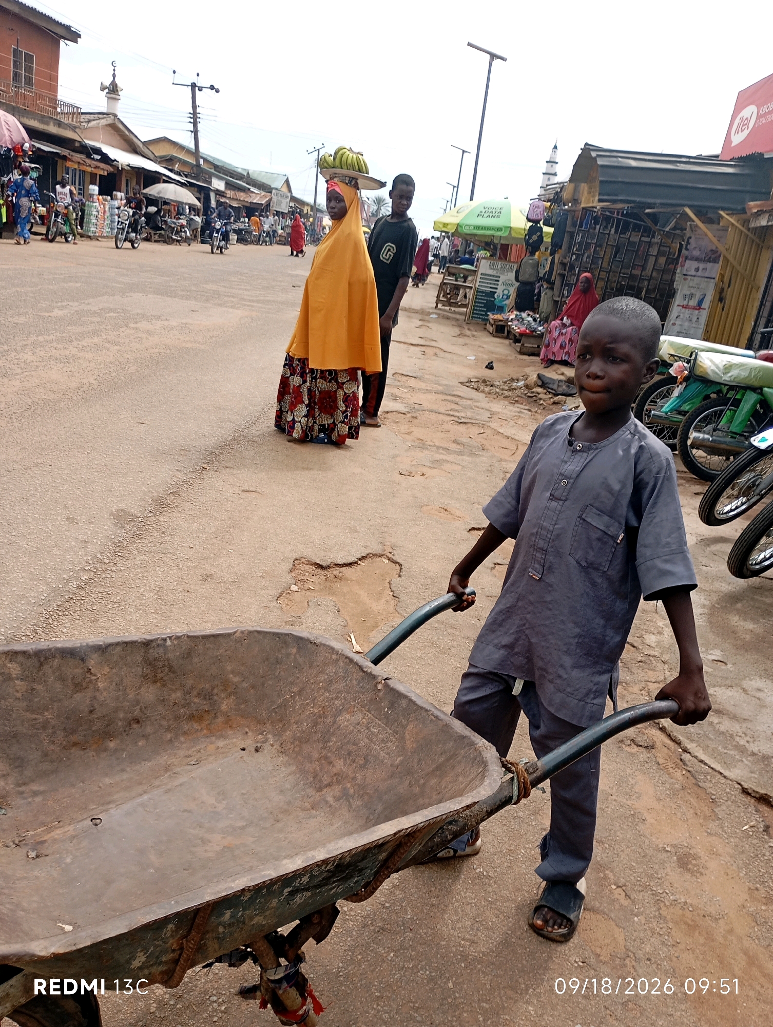 The image depicts a street scene in Shendam, Nigeria, focusing on a young boy pushing a wheelbarrow. The boy, wearing a gray tunic and pants, is in the foreground, holding the handles of the weathered, metal wheelbarrow. Behind him, a woman in an orange headscarf and a patterned skirt stands with a tray of bananas on her head. Further in the background, a bustling market environment is visible, with stalls, motorcycles, and other pedestrians. Buildings line the street, and a mosque minaret can be seen in the distance. The ground is paved with asphalt and dirt. The overall impression is one of daily life and commerce in a Nigerian town.