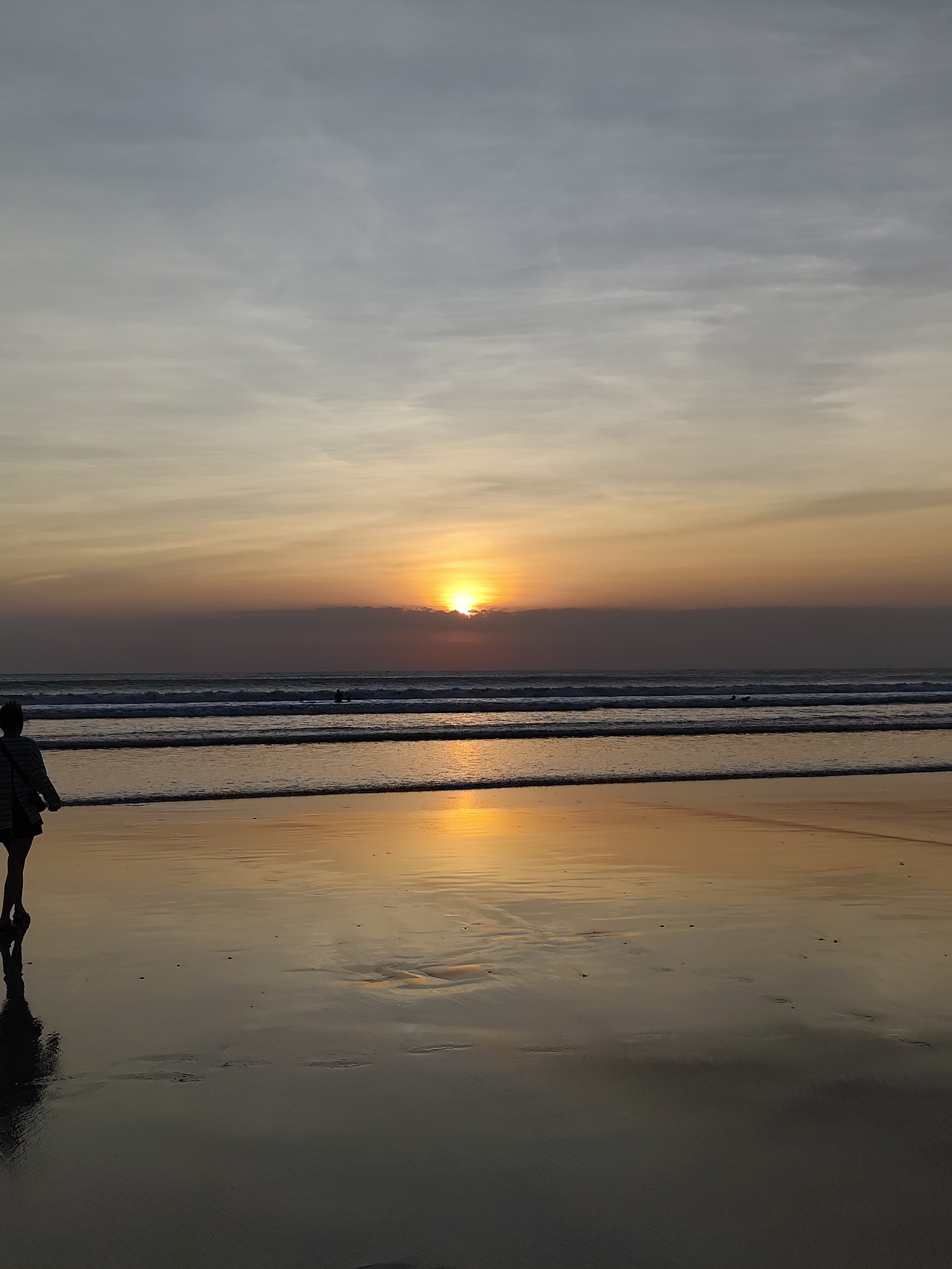 A person walks on a sandy beach towards the ocean at sunset. The sun is low on the horizon, casting a warm glow on the water and the wet sand, creating a reflective surface. Waves gently lap at the shore. The sky is a gradient of pale blue and orange, with scattered clouds. In the distance, a few surfers can be seen in the water. The scene is tranquil and serene. The location is unknown.