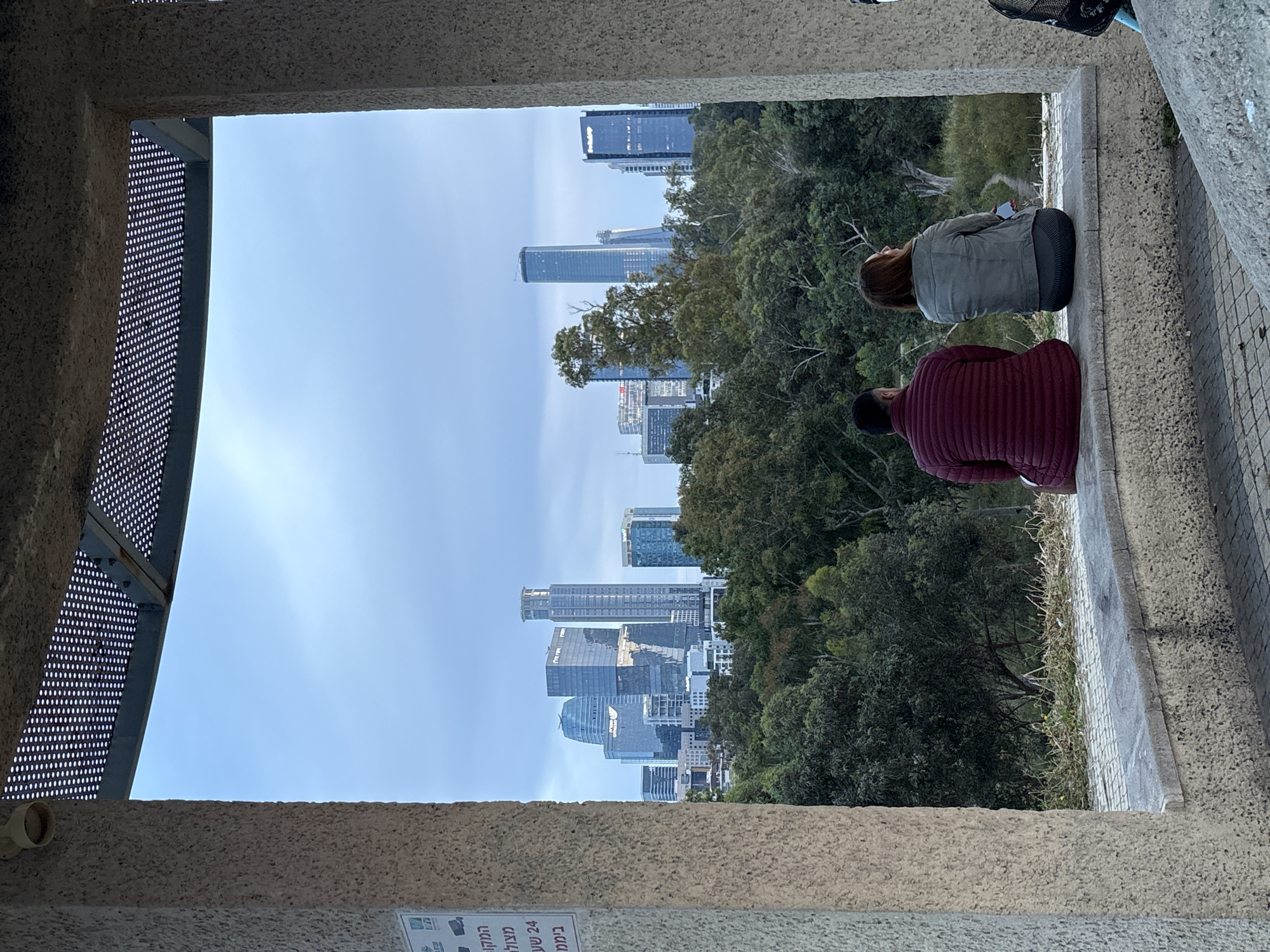 The image, when correctly oriented, captures a framed view from what appears to be a lookout point or balcony. The foreground is dominated by a rough-textured concrete structure, with a perforated metal panel visible on the left side, acting as part of the frame.
Two individuals are seated on a concrete ledge or bench in the mid-ground, seen from behind. One wears a gray jacket, and the other is dressed in a burgundy puffer jacket. They appear to be observing the panorama before them.
Beyond the people, a lush green area with trees and dense foliage transitions into a modern cityscape. Numerous high-rise buildings and skyscrapers punctuate the horizon under a clear, bright blue sky with faint, wispy clouds, suggesting a daytime scene. A bicycle seat and part of a basket are partially visible in the upper right corner, indicating a bike is nearby.
A notable detail is a sign with Hebrew text affixed to the concrete wall in the lower-left corner. The text reads "המקום מצולם 24 שעות ביממה" which translates to "The place is filmed 24 hours a day," indicating surveillance. The setting combines natural greenery with a bustling urban skyline, offering a tranquil spot for observation in Tel Aviv, Israel. The activity appears to be a moment of relaxation or contemplation.