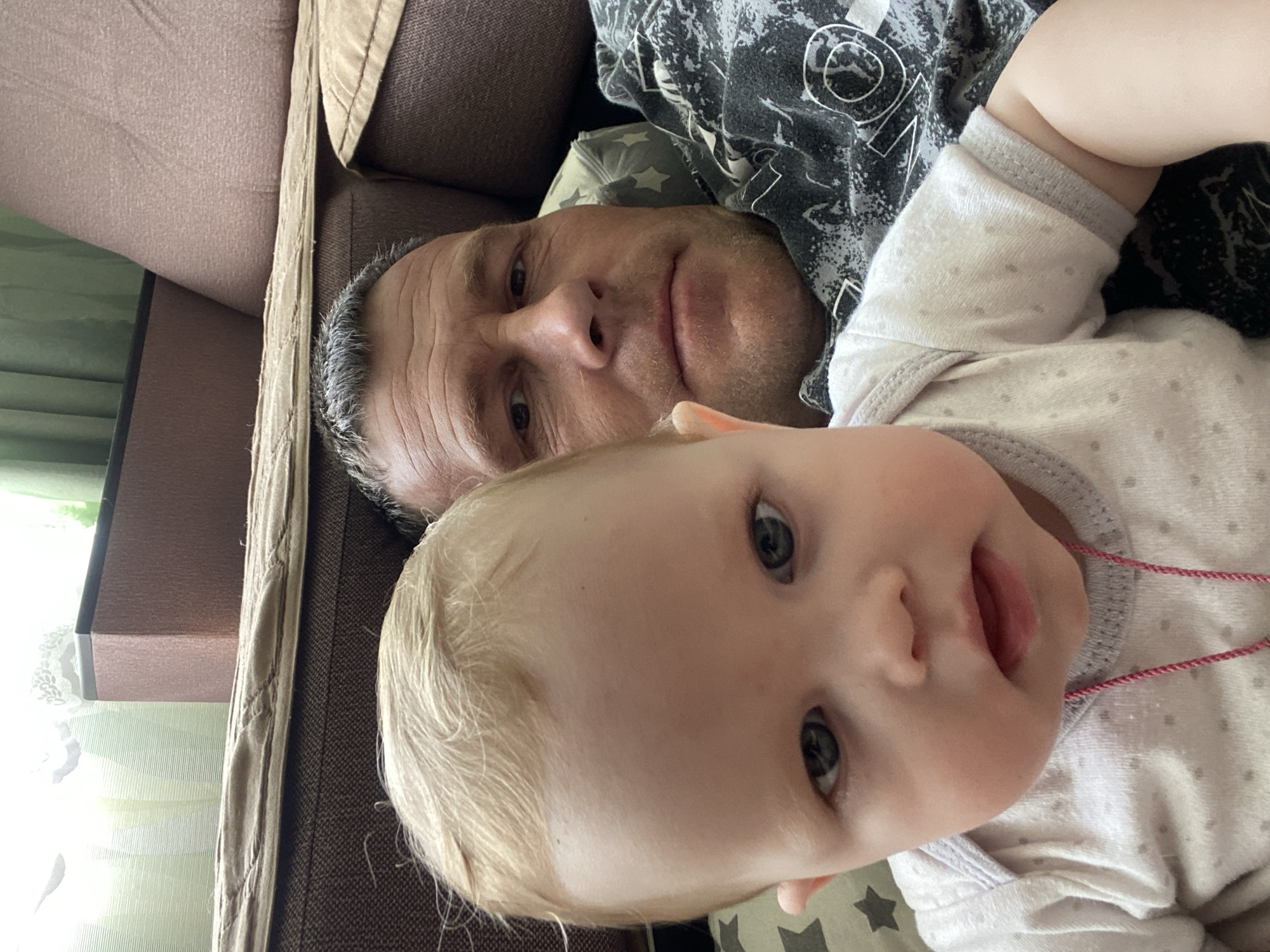 A close-up, canted angle shot shows a man and a baby lying down together indoors. The man is positioned behind the baby, looking at the camera with a slight smile. He has short, graying hair and is wearing a dark-colored shirt with a patterned design. The baby is in the foreground, also looking at the camera. The baby has fair skin, blonde hair, and is wearing a light-colored onesie with a pattern of small gray dots. A red string is visible near the baby's neck. The background includes part of a brown couch and a curtain. No information about the location of Фоки, Russia is present in the image.