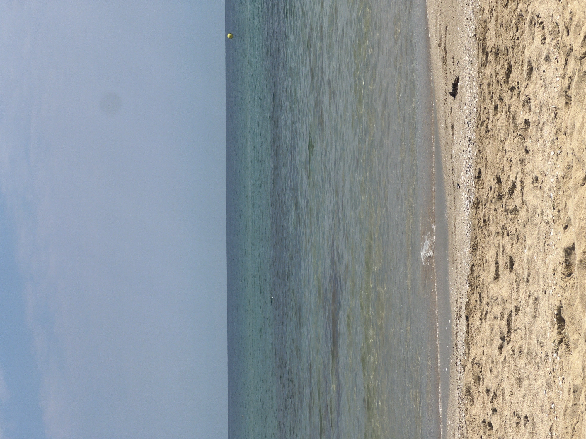 The image depicts a tranquil seascape with a focus on the beach and the ocean. The foreground is dominated by a sandy beach, showing footprints and scattered seashells. The water is clear and shallow near the shore, gradually deepening and changing to a deeper blue-green hue further out. The horizon is clearly defined, separating the sea from a pale blue sky with a few faint clouds. A single yellow buoy is visible in the distance on the right side of the ocean. The scene conveys a sense of calm and natural beauty, likely a section of the coastline near Sozopol, Bulgaria.