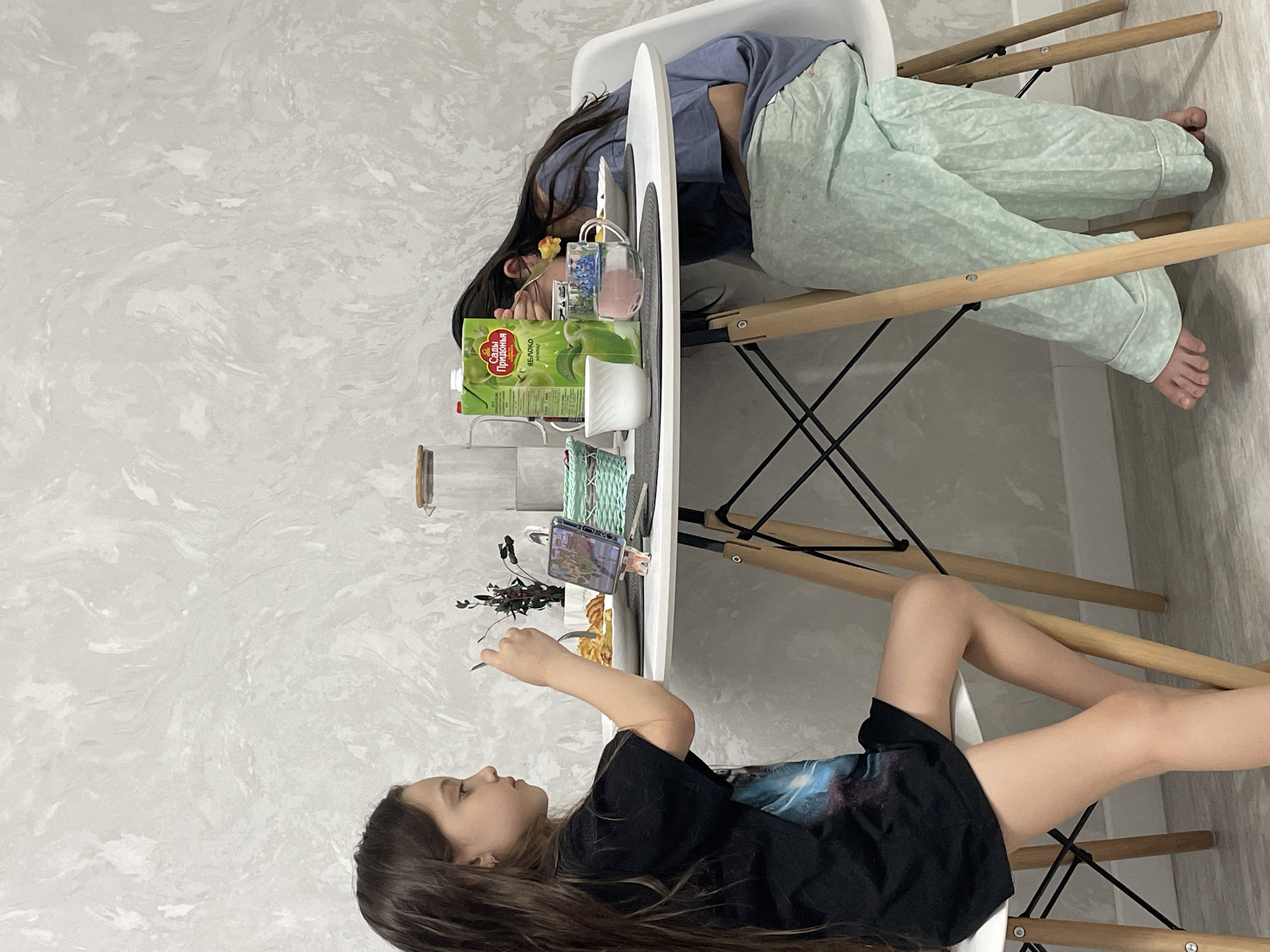 Two girls are seated at a white table in a room with light gray walls. One girl is resting her head on the table, clad in a light blue shirt and light green patterned pants. A box of apple juice, a cup, and other items are on the table. The second girl sits across from her, dressed in a black t-shirt and shorts, looking at a phone propped up on the table. The legs of the table and chairs are wooden with black metal supports. The scene takes place in a domestic setting. No information about Izhevsk, Russia is evident.