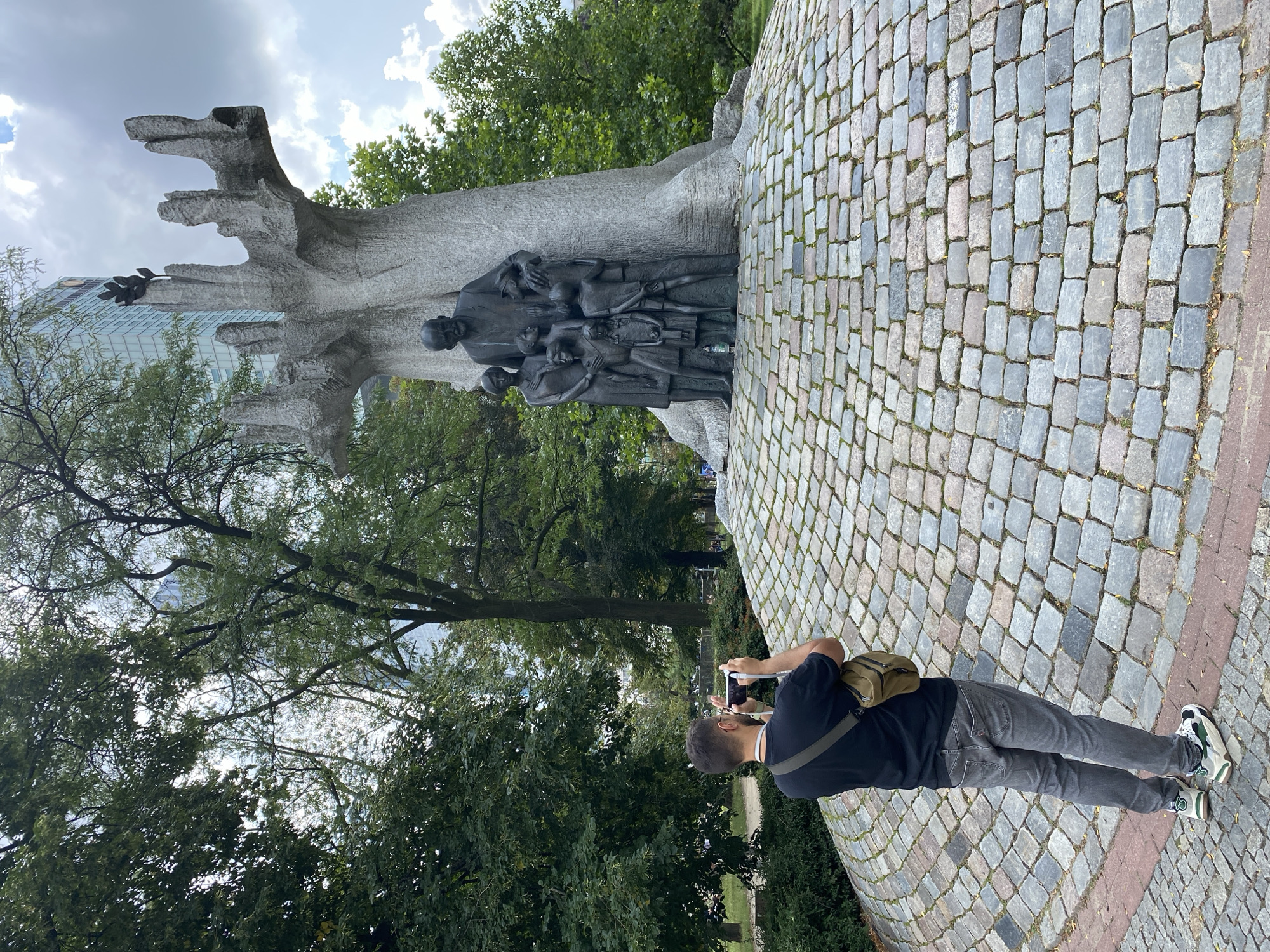 In Warsaw, Poland, a man is seen photographing a monument of a man embracing a group of children. The monument is made of stone and is situated on a cobblestone pedestal surrounded by trees and a modern skyscraper in the background. The man is wearing a black t-shirt, grey jeans, and a crossbody bag. He is holding a smartphone in his hands, aiming it at the monument. The sky is overcast with some clouds.