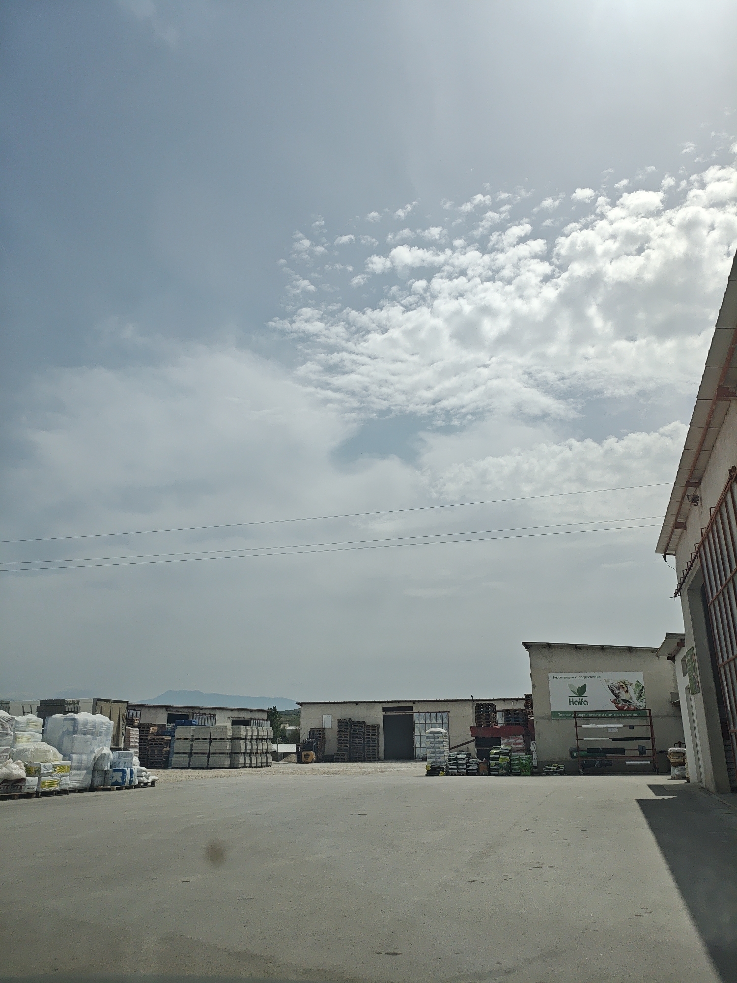 The image shows an outdoor industrial area with various materials and structures. In the foreground is a large paved area with some dirt or stains visible. To the left, there are stacks of bags and wrapped goods, indicating a storage or distribution facility. Further back, there are pallets stacked with tiles or concrete blocks. Buildings with multiple entry points line the background, and a billboard with the word "Haifa" is present. Electrical wires stretch across the sky, which is partly cloudy. In the distance, there are faint hills or mountains. A portion of a building or structure with a red-colored element is visible on the right side of the frame.