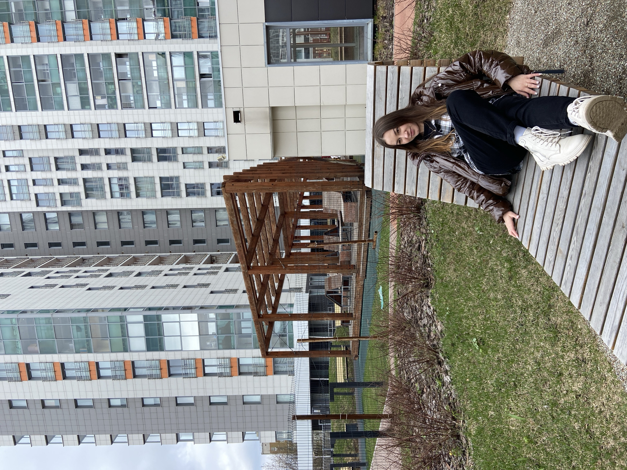 A young woman is casually seated on a wooden slatted bench in an outdoor urban environment in Izhevsk, Russia. The original image is rotated 90 degrees counter-clockwise. She has long brown hair and is wearing a shiny brown puffer jacket, a plaid shirt, black pants, and light-colored high-top boots with white socks. She looks towards the camera with a relaxed, slight smile.
The bench is situated on a ground area that combines sparse green grass with gravel. Behind the woman and the bench, a chain-link fence separates the foreground from a larger grassy expanse and a prominent wooden pergola structure with an open framework. In the background, towering modern apartment buildings dominate the scene. These multi-story structures feature numerous windows of various sizes and arrangements, some with distinct orange accents or dark frames, set against light-colored and grey facade panels. The overall setting suggests a residential area, with bare bushes flanking the grassy areas, indicating a late winter or early spring season. The lighting is natural and appears to be from an overcast or partly cloudy daytime sky. No visible text is present in the image.