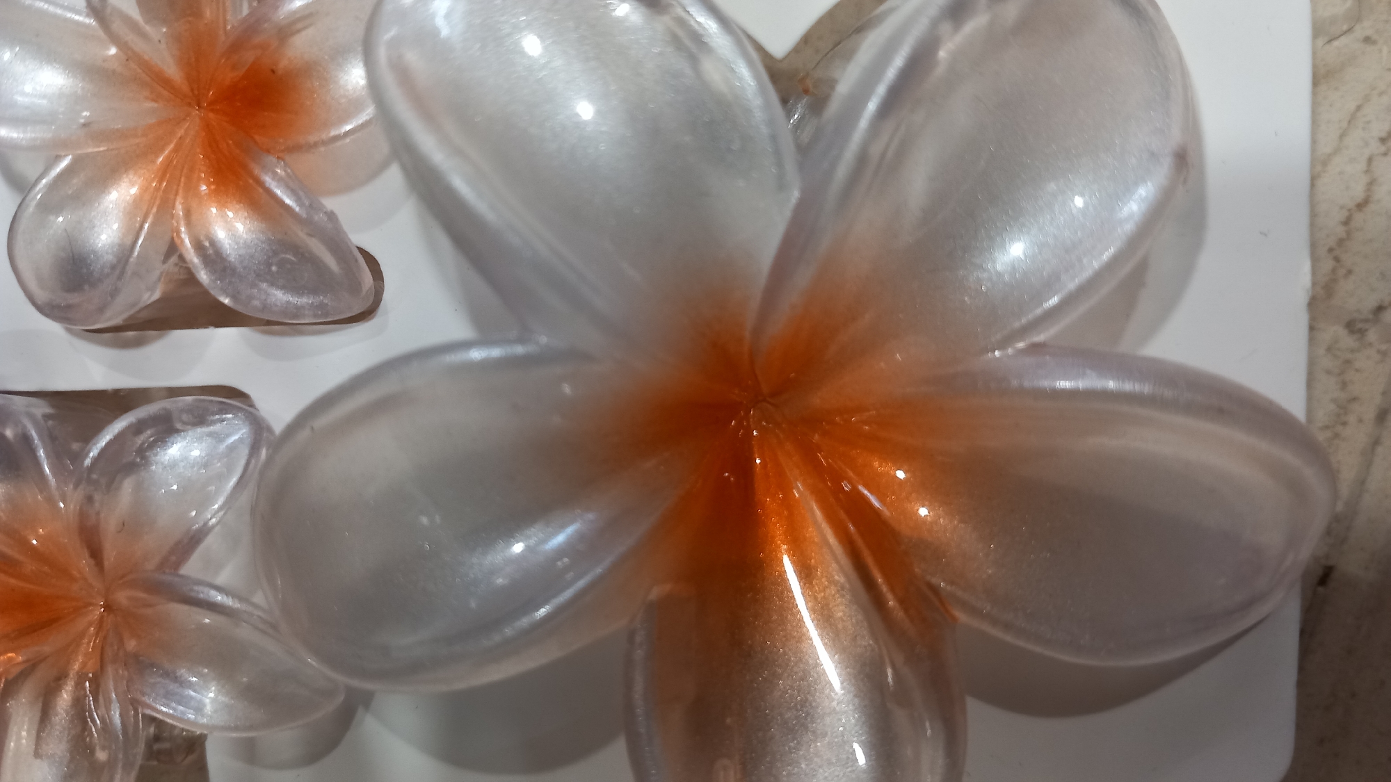 The image shows a close-up of several clear, translucent, plastic decorative flowers with iridescent, orange centers. The flowers have five petals each, and their surfaces are smooth and shiny, reflecting light. The flowers appear to be arranged on a flat, light-colored surface, possibly a table or shelf. The background is out of focus but suggests an indoor setting. There are no people or animals visible in the image. The overall impression is one of delicate, decorative objects. There is no information in the image that suggests a specific location in Kos, Greece.