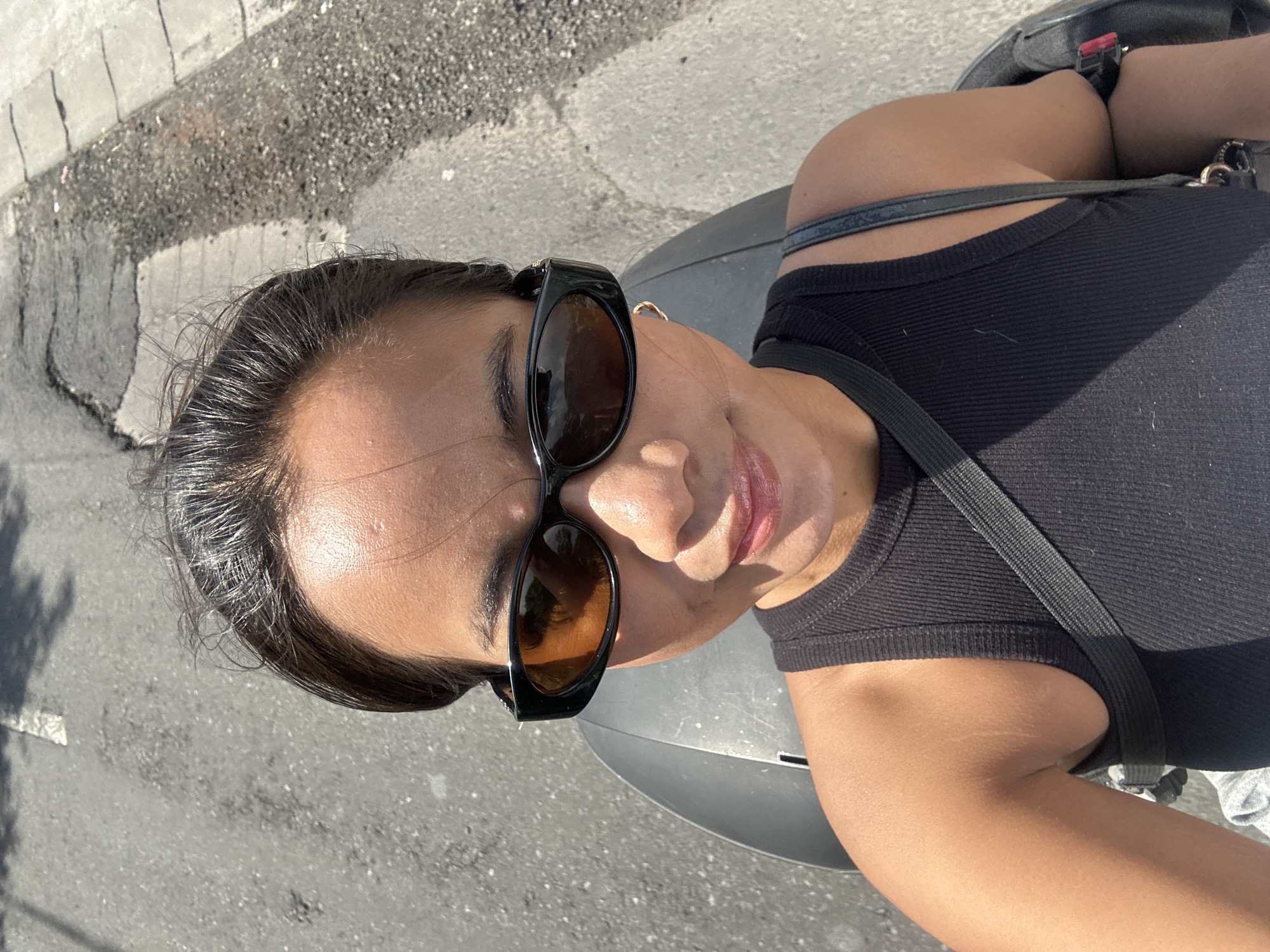 A woman with dark hair pulled back is taking a selfie outdoors. She is wearing sunglasses with orange lenses and a black sleeveless top. A gray strap is visible across her chest. The background shows a cracked asphalt road with some white markings, suggesting an urban or semi-urban environment, possibly in Sukawati, Indonesia. The sunlight indicates it is daytime.