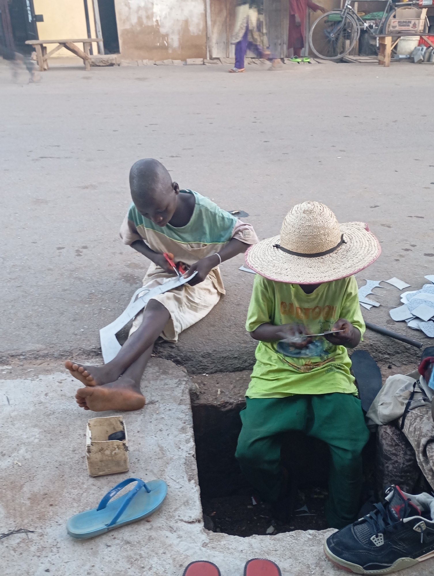 The image depicts two young boys working on a street in an outdoor market environment. The boy on the left is seated on a curb, wearing a light-colored shirt with green accents, and is using red-handled scissors to cut a pattern out of white paper. His legs are crossed, and his feet are bare. To his right, another boy is seated, wearing a large straw hat, a green t-shirt with a graphic design, and green pants. He appears to be holding something in his hands, possibly working with a similar material to the boy on the left. Scattered around the boys are various pieces of cut paper or leather, suggesting they are in the process of crafting something, likely footwear. A blue flip-flop and a small white box with a dark object inside are visible on the ground near the boy on the left. In the background, there is a dirt road or paved street with some buildings and a bicycle. The scene suggests a resourceful and industrious environment, possibly indicating child labor or a traditional craft being practiced.