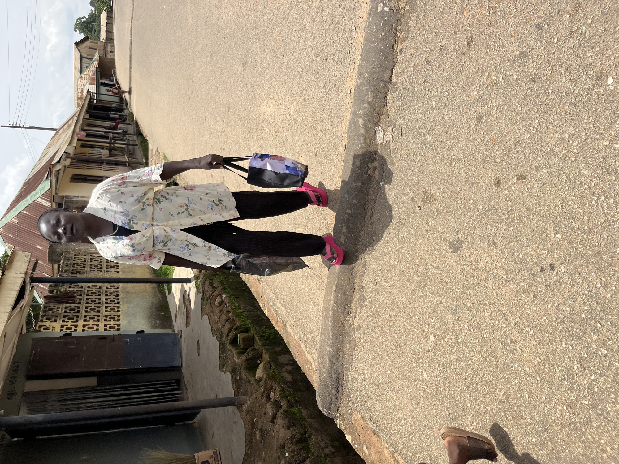In Bukuru, Nigeria, a woman with dark skin walks towards the viewer on an asphalt road. She wears a light-colored floral print blouse, black pinstripe pants, and bright pink flip-flops. She carries a black plastic bag in her left hand and a patterned tote bag in her right. Behind her, to the left, is a building with a corrugated metal roof and decorative concrete blocks. To her right, the road stretches into the distance, lined with similar buildings and a few scattered pedestrians. The sky is overcast.