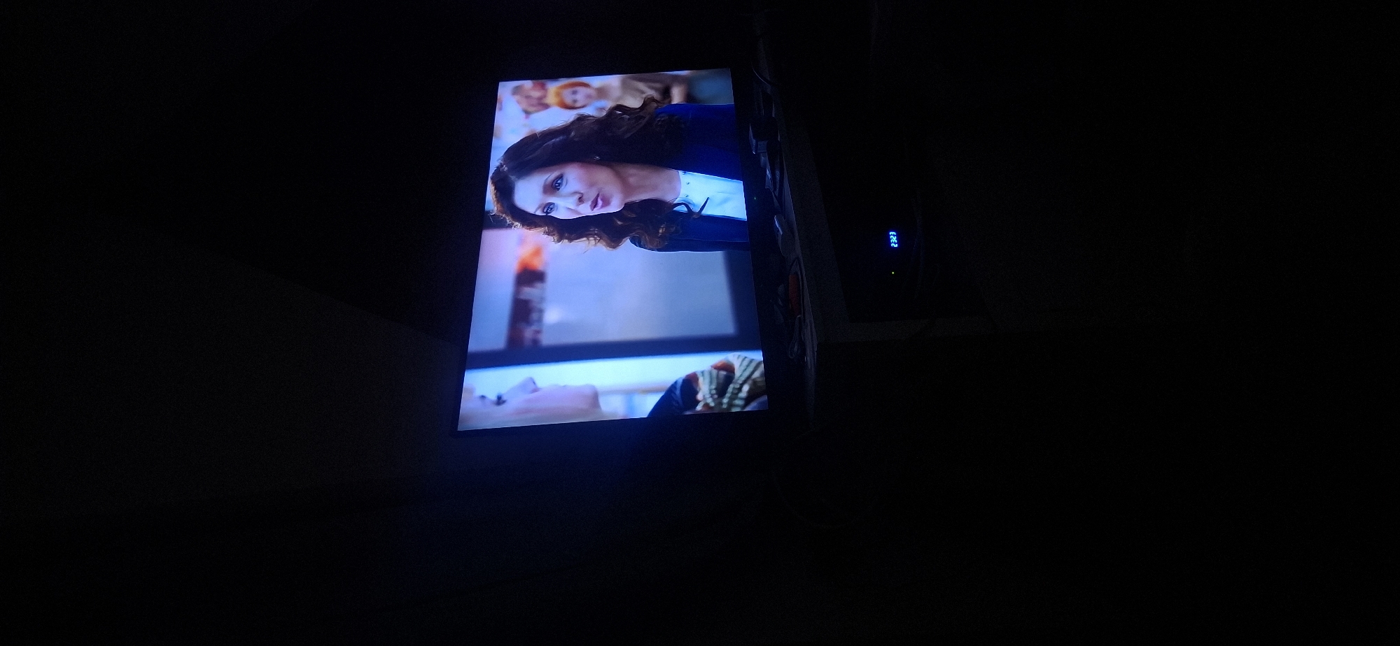 The image shows a television screen displaying a scene from a show or movie. On the screen, there are two women. The woman on the right has dark, wavy hair and is wearing a dark blue jacket over a white top. She is speaking. To her left, partially visible, is another woman with blonde hair. In the background, behind the women, is a display case with items that are difficult to discern. Below the television, on a dark surface that appears to be a stand or entertainment center, a digital clock displays "23:23". The overall environment is dark, suggesting the picture was taken in a dimly lit room. The scene is not located in Yerocham, Israel.