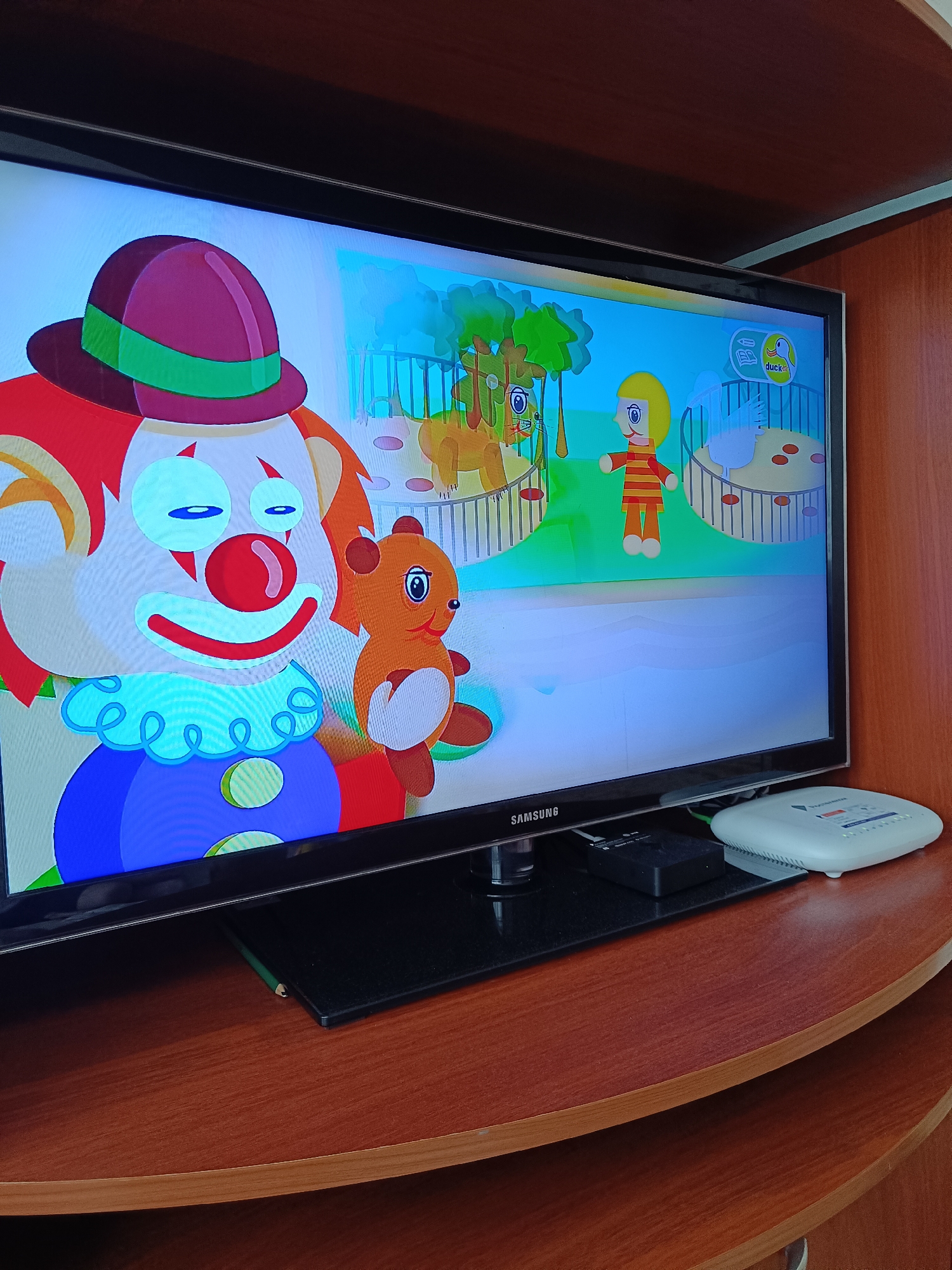 A Samsung television displays a cartoon featuring a clown and a bear. The television is situated on a wooden shelf, accompanied by a small black device and a white modem. The image was taken in an indoor setting. The location of the city of Tobolsk, Russia, cannot be confirmed.