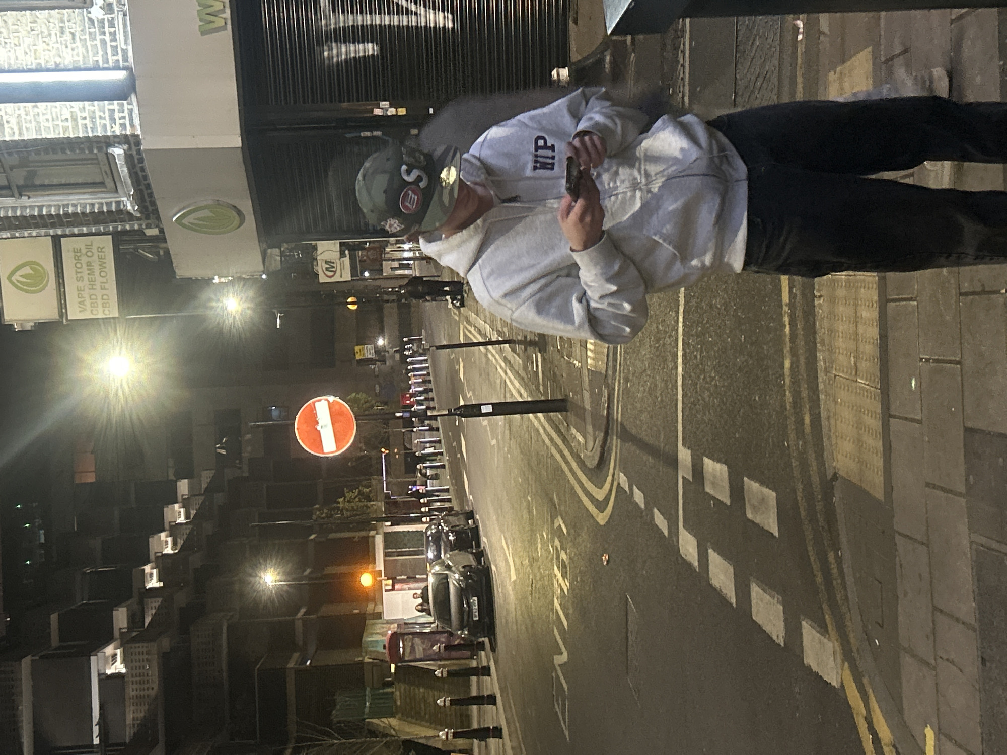 A street scene at night in London, United Kingdom. A person stands on the sidewalk looking at a phone. The person wears a grey hoodie with "HIP" written on it, a cap, and dark pants. To the left is a building with a sign indicating a vape store selling CBD hemp oil and CBD flower. A street with painted markings runs along the right side of the image, with parked cars visible in the distance. A "No Entry" sign is also visible. The scene is illuminated by streetlights. The architectural style of the buildings in the background appears consistent with the area.