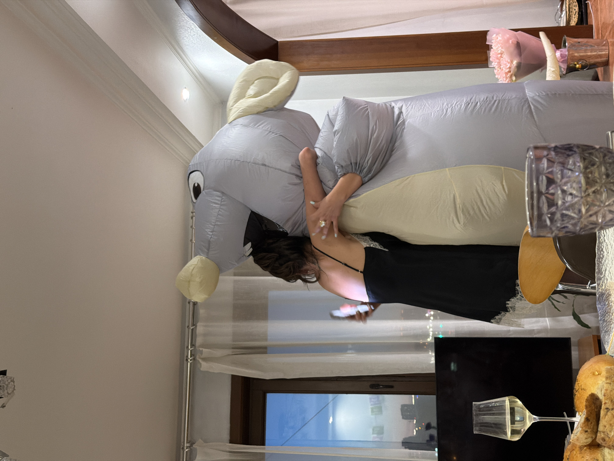 Here is a description of the image:
A woman is wearing an inflatable elephant costume inside what appears to be a living room. The room contains a window with sheer curtains, a television, a glass, and some bread. The woman is holding a mobile phone in her hands. The location is Unknown, Unknown.