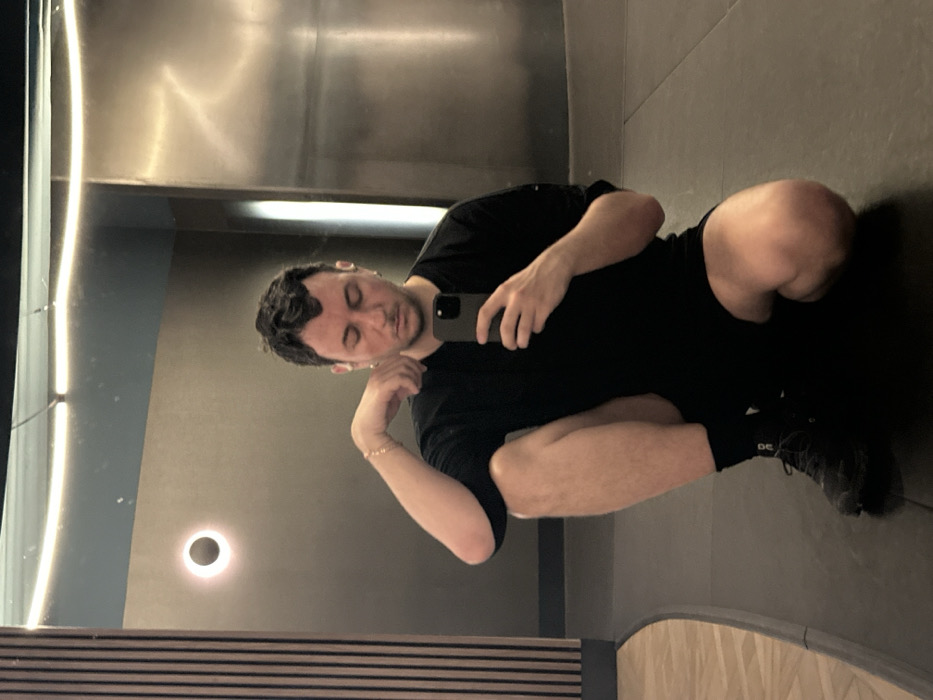 A male individual is depicted in a full-length mirror selfie, crouching on a dark floor within a modern interior space. The subject, with dark wavy hair, is wearing a black short-sleeved t-shirt, black athletic shorts, black socks, and black athletic shoes. A light-colored chain bracelet is visible on his left wrist and small earrings in both ears. He holds a dark-colored smartphone in his right hand, angled towards himself, with his gaze directed downwards. His left arm rests on his left thigh, hand positioned near his left ear/neck.
The environment, visible through reflection, features dark walls. A continuous horizontal LED strip light illuminates the upper section of the mirror. To the left, a circular wall sconce emits a glowing halo effect, and vertical ribbed wooden paneling is visible. The dark floor where the individual squats transitions with a curved edge to a lighter, possibly parquet, floor section further left. A metallic or polished panel with a vertical light reflection is present on the right. A section of dark blue/teal colored ceiling or wall is discernible in the upper left corner.
The exact location within London, United Kingdom, is not discernible from the visual information; however, the contemporary interior design and features are consistent with a modern fitness facility, such as a high-end gym or a hotel's exercise area.