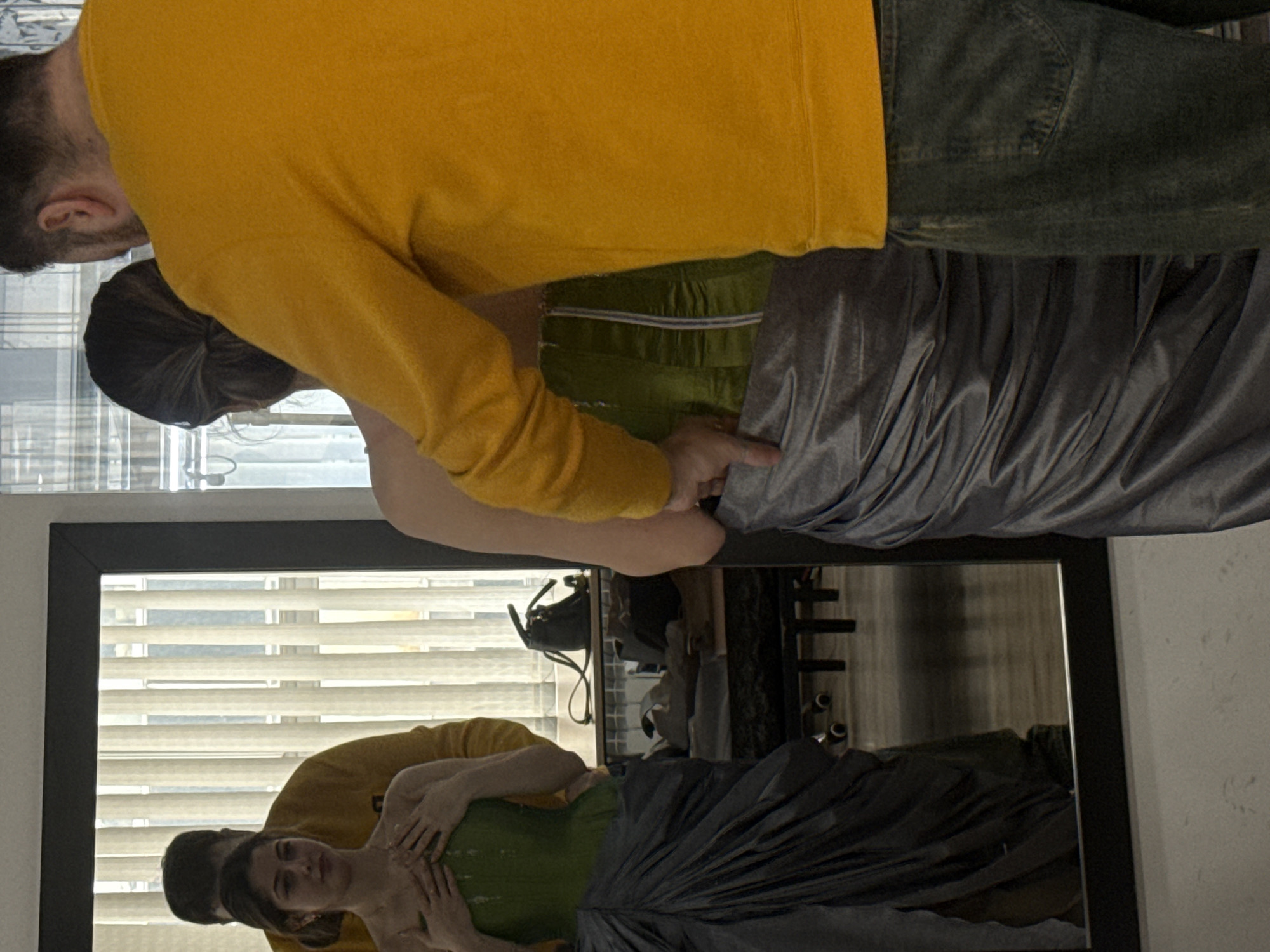 A woman in a green dress and grey fabric is being helped by a man in a yellow sweatshirt and jeans in what appears to be an indoor setting, with their reflections visible in a black-framed mirror. Blinds are seen in the background, and a purse is hanging in the mirror's reflection. The scene is not conclusively located in Yerevan, Armenia, based solely on the provided image.